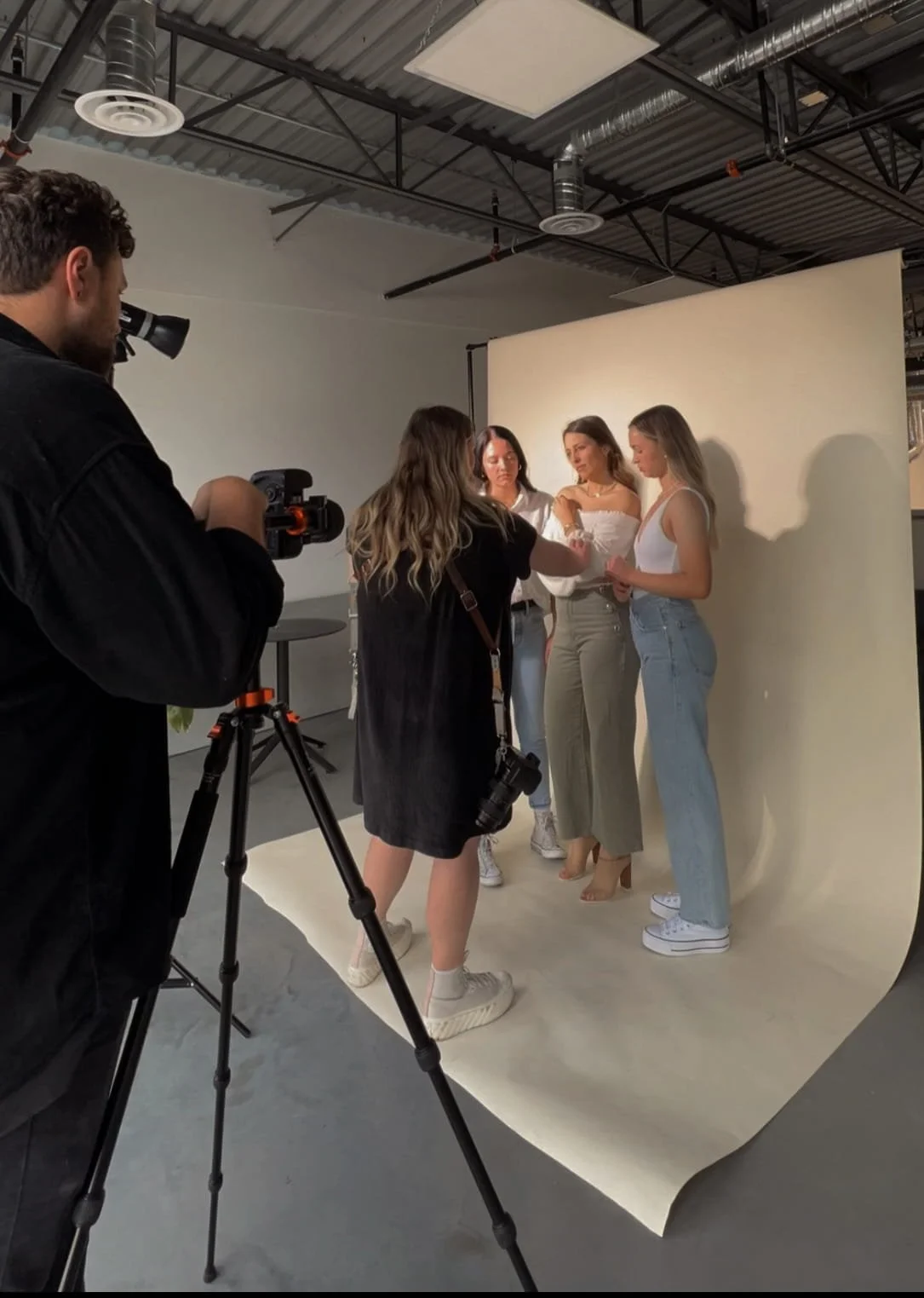 A behind-the-scenes brand content shoot with a photographer, videographer, and three women posing against a neutral backdrop in a professional studio setting.