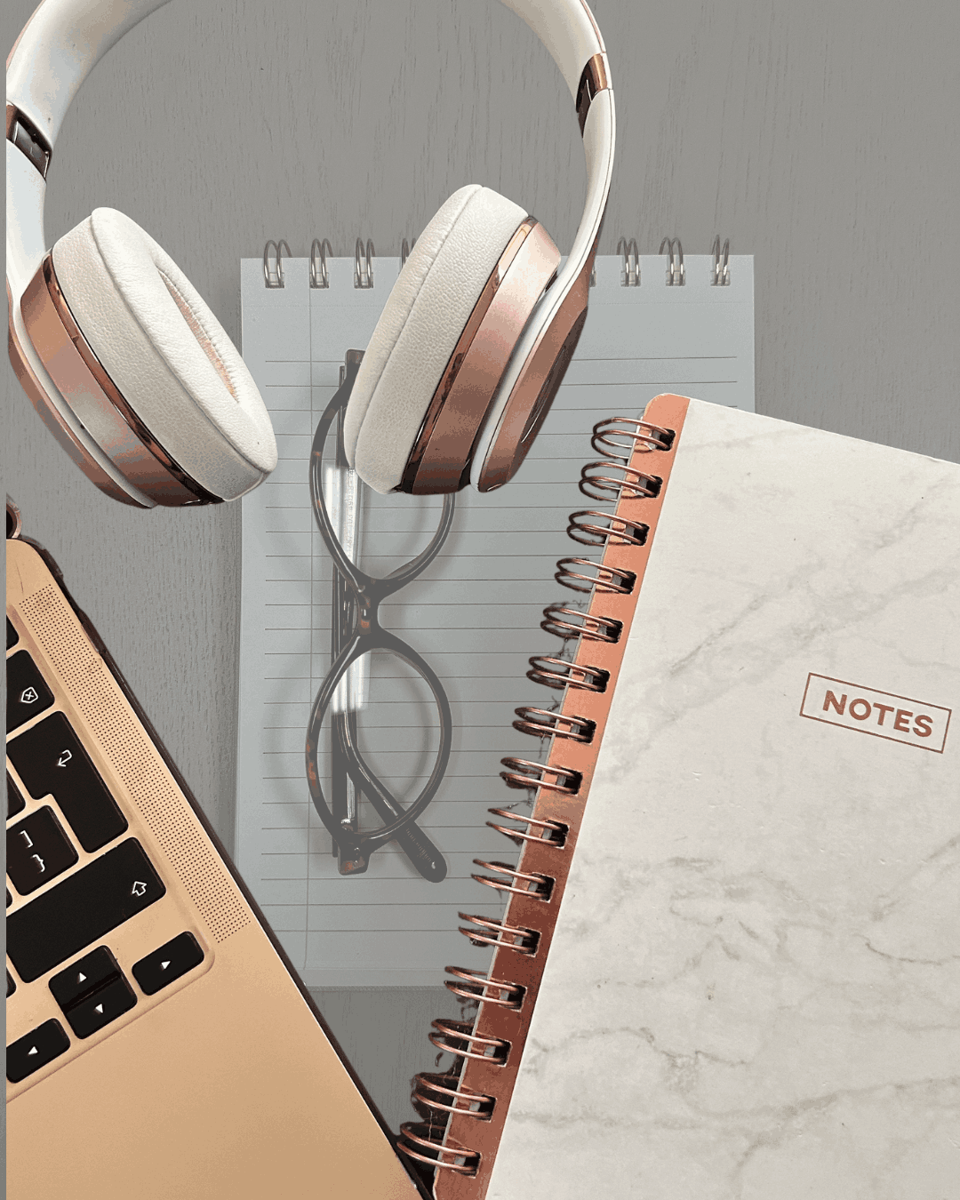 Flat lay of a rose gold laptop, headphones, glasses, and marble notebook labeled 'Notes' on a desk, representing a workspace for brainstorming a marketing brief.