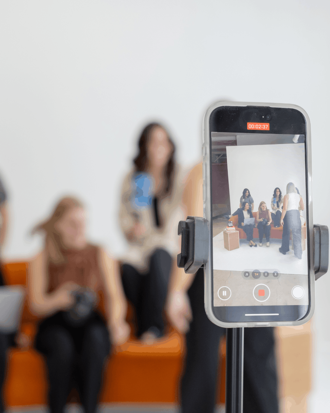 iPhone mounted on a tripod actively recording a group of women seated and standing in a studio, with the recording screen visible and subjects blurred in the background