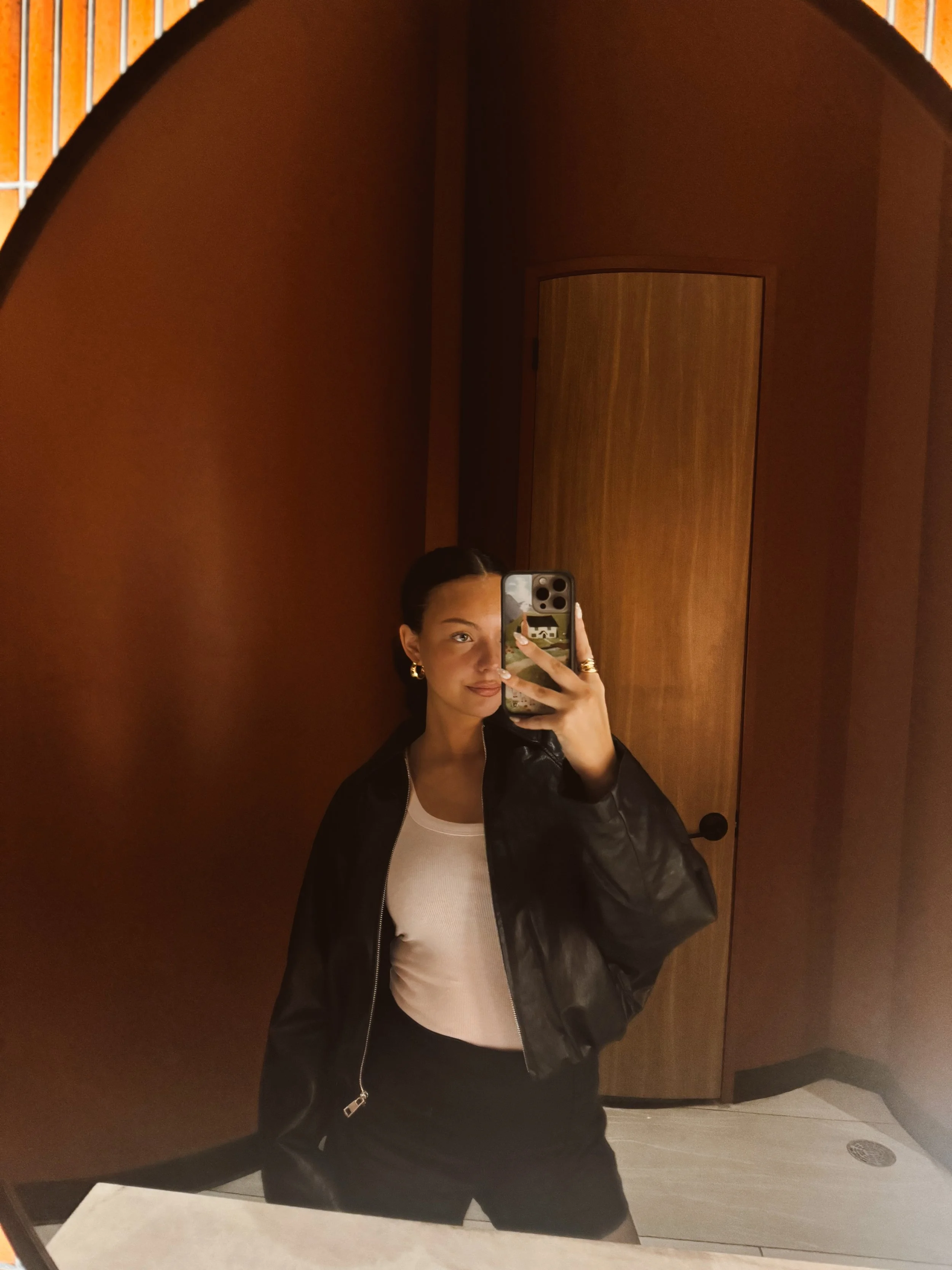 Mirror selfie featuring a black oversized bomber jacket, white tank, and gold hoop earrings against a warm-toned arched mirror.