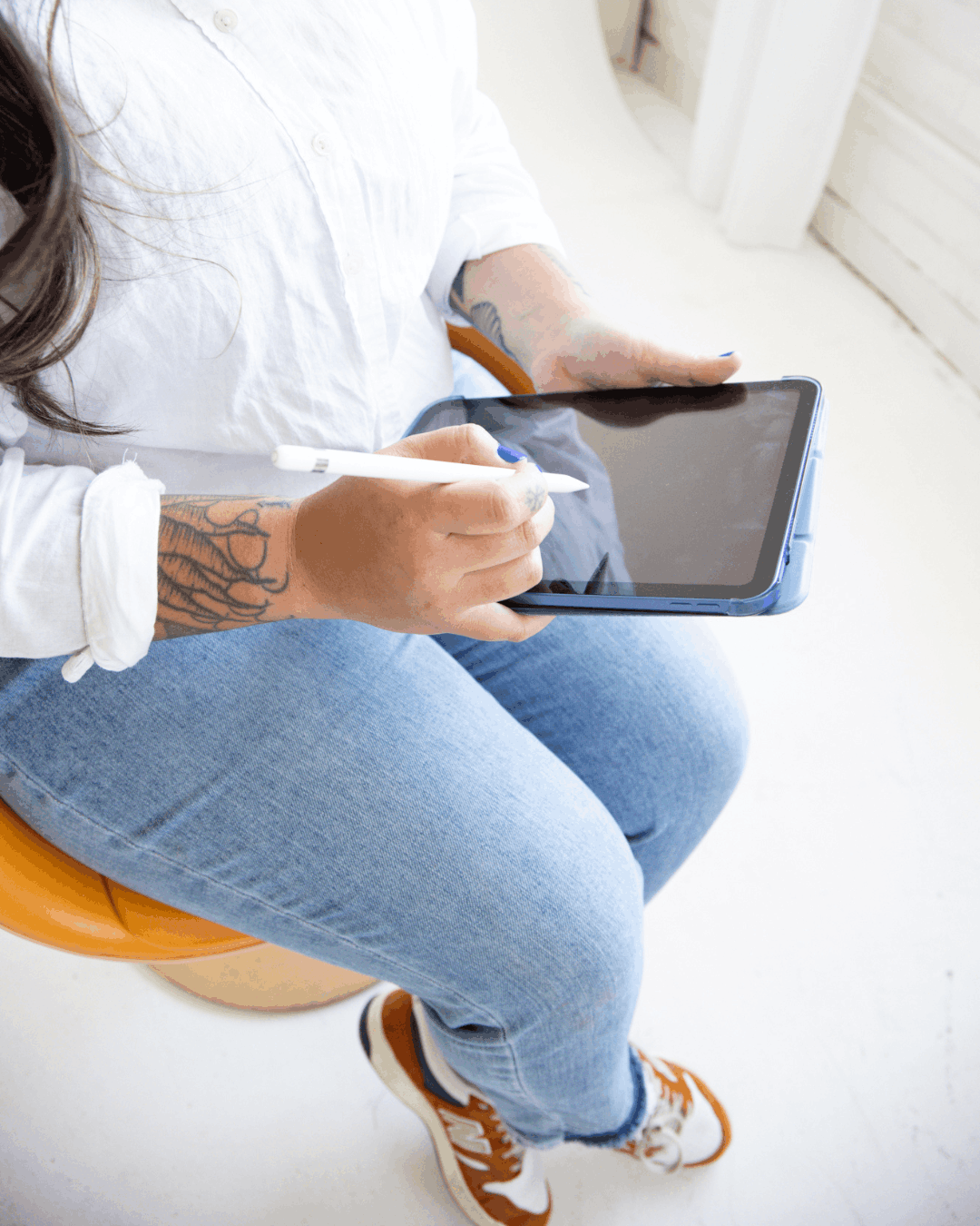 A tattooed woman sitting and holding an iPad with an Apple Pencil, planning or sketching content strategy in a bright, minimal workspace.