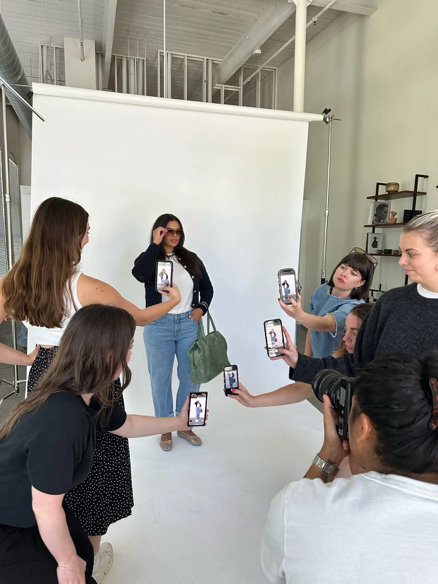 Behind-the-scenes of a photo shoot: a model poses in front of a white seamless backdrop while multiple photographers and crew members photograph her with phones and a DSLR camera in a bright studio space.