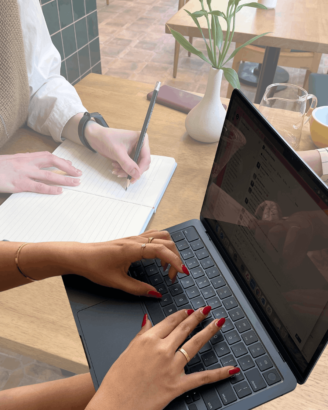 Two people collaborating at a café table, one writing notes by hand and the other typing on a laptop, representing a strategy partnership discussion."
