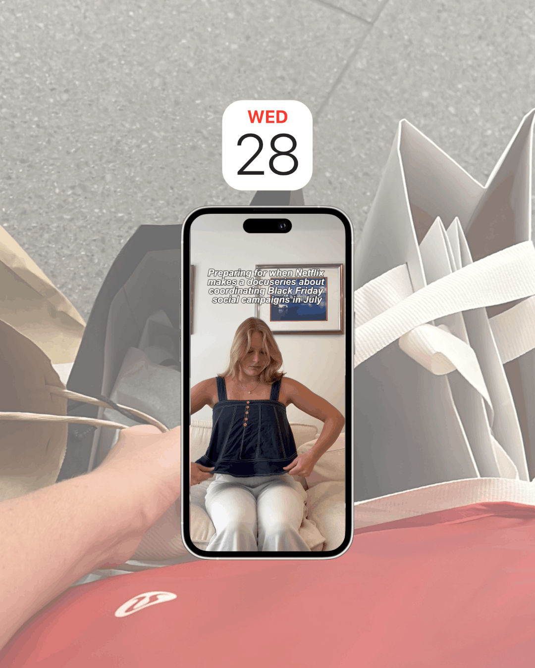 Woman adjusting outfit in a phone-screen video with caption about coordinating Black Friday social campaigns in July, set against a background of shopping bags and a 'Wed 28' calendar reminder.