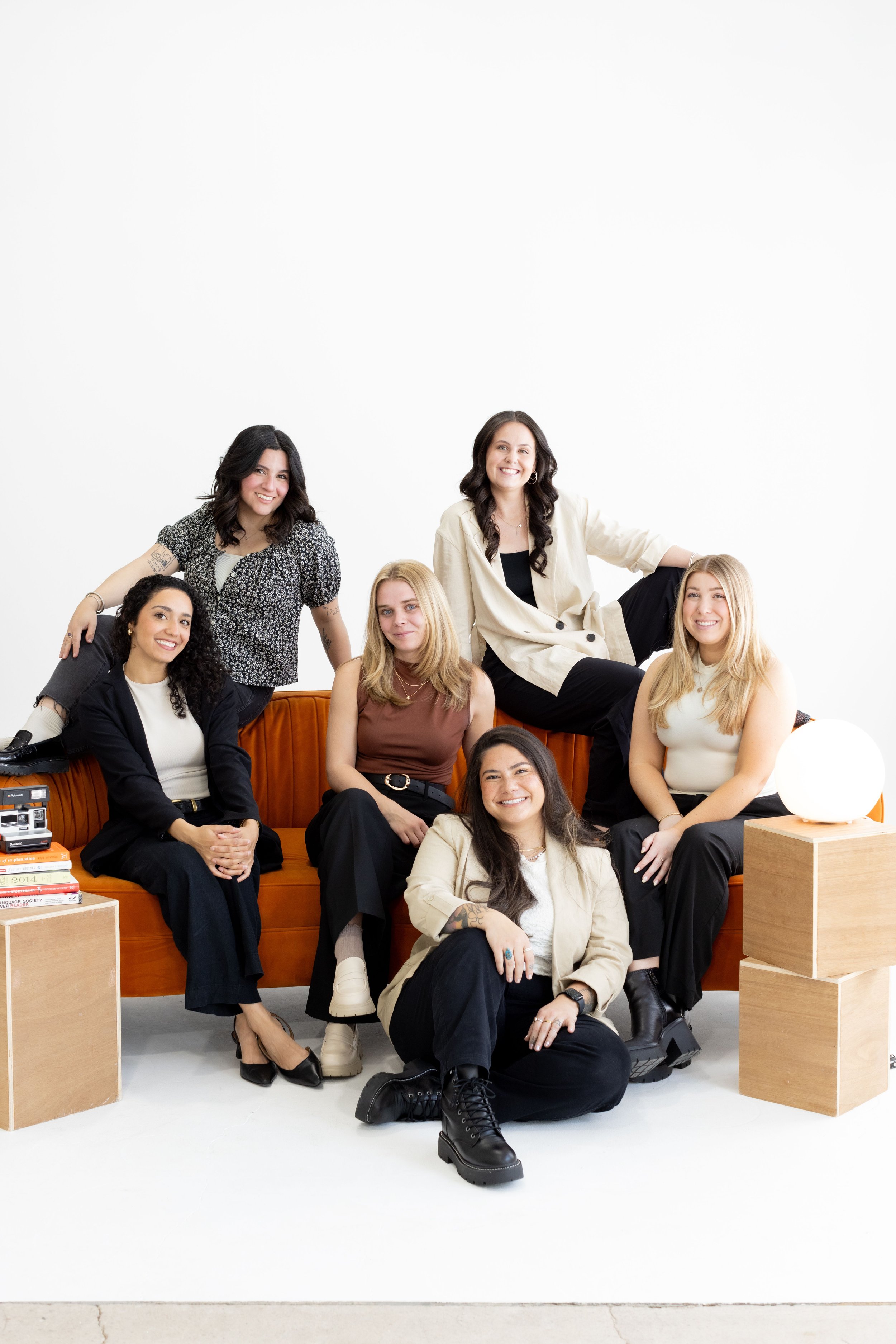 Group of seven women sitting and standing on an orange sofa in a studio with a plain white background, arranged in a casual pose, smiling and dressed in stylish casual clothing.
