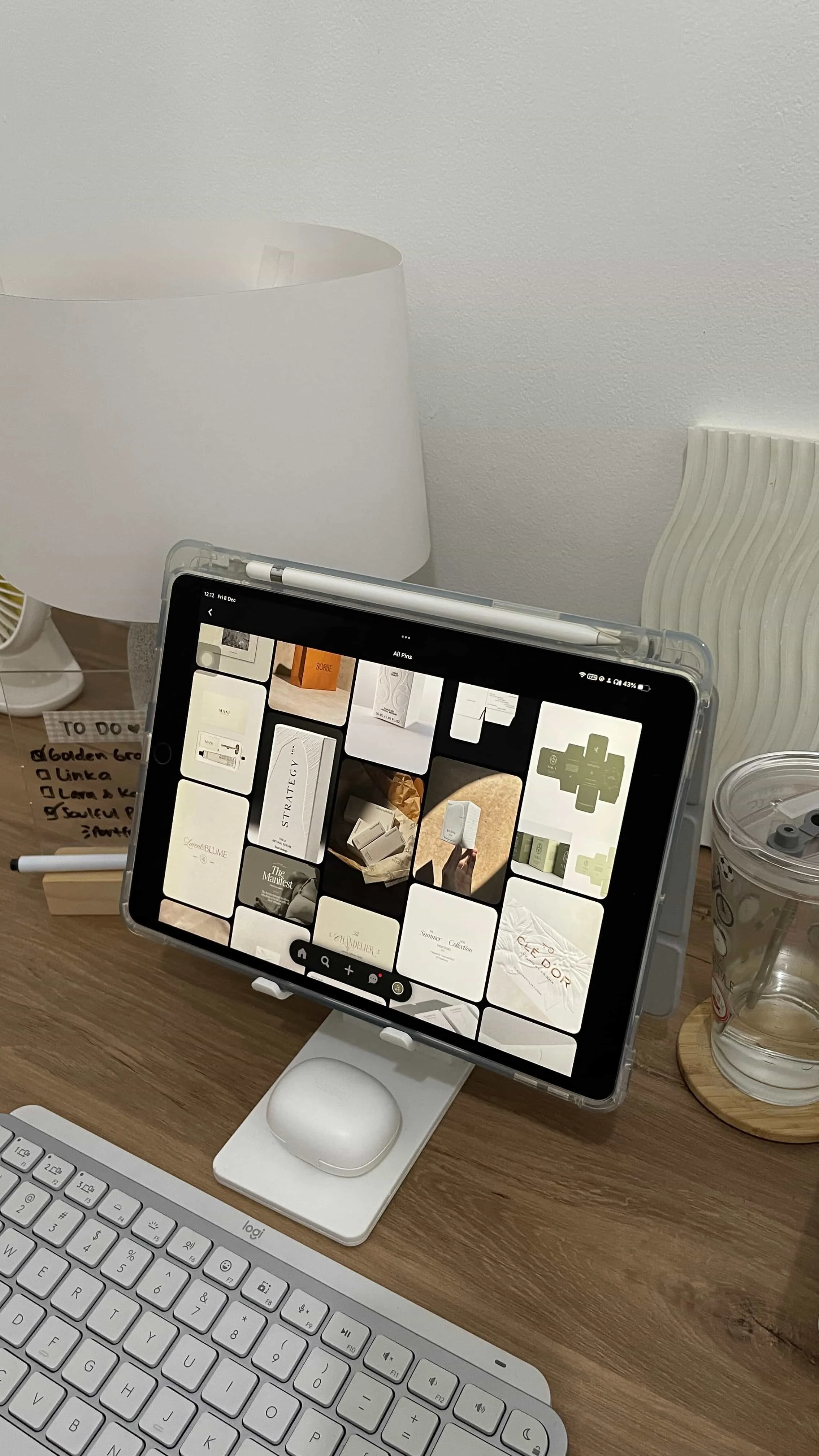 iPad on a desk stand displaying a Pinterest mood board of neutral, minimalist brand design inspiration, surrounded by a Logitech keyboard, Apple Pencil, AirPods case, and a handwritten to-do list