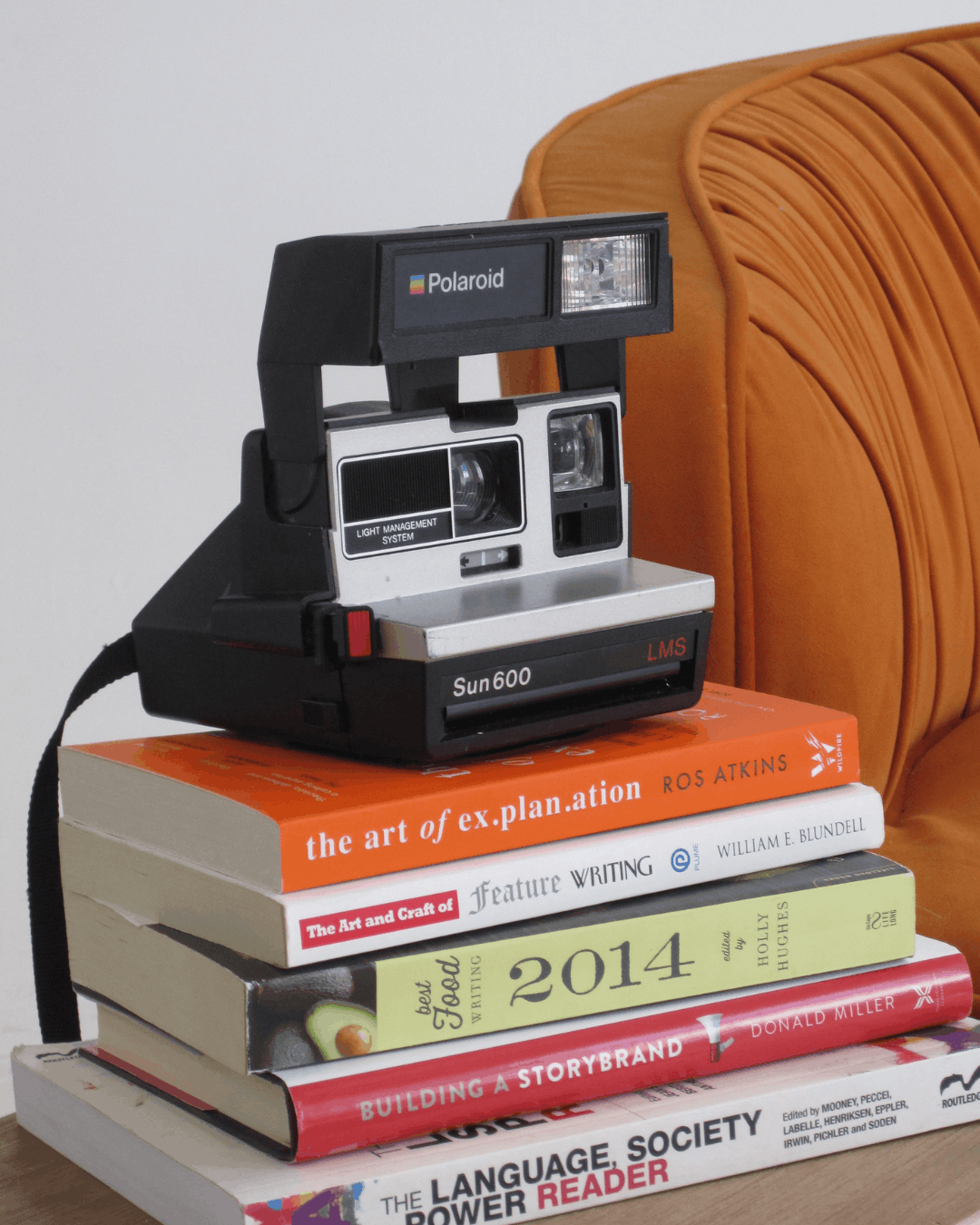 A vintage Polaroid Sun 600 camera resting on a stack of marketing and writing books including Building a StoryBrand and The Art of Explanation, styled against an orange chair for a creative agency flat lay.