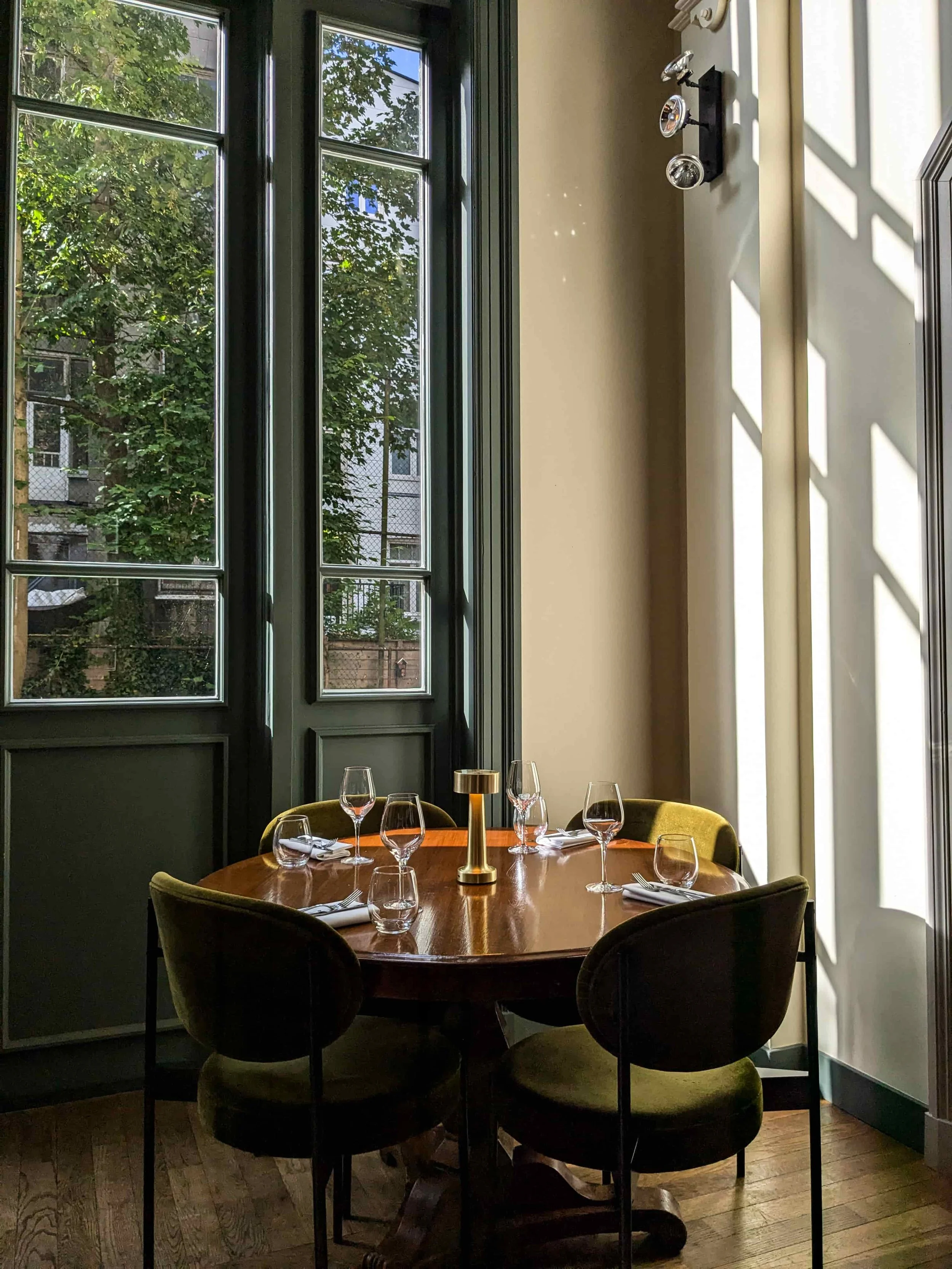 An elegant restaurant dining table set with wine glasses and a brass candle lamp, surrounded by olive green velvet chairs, beside tall dark-framed windows overlooking leafy trees.