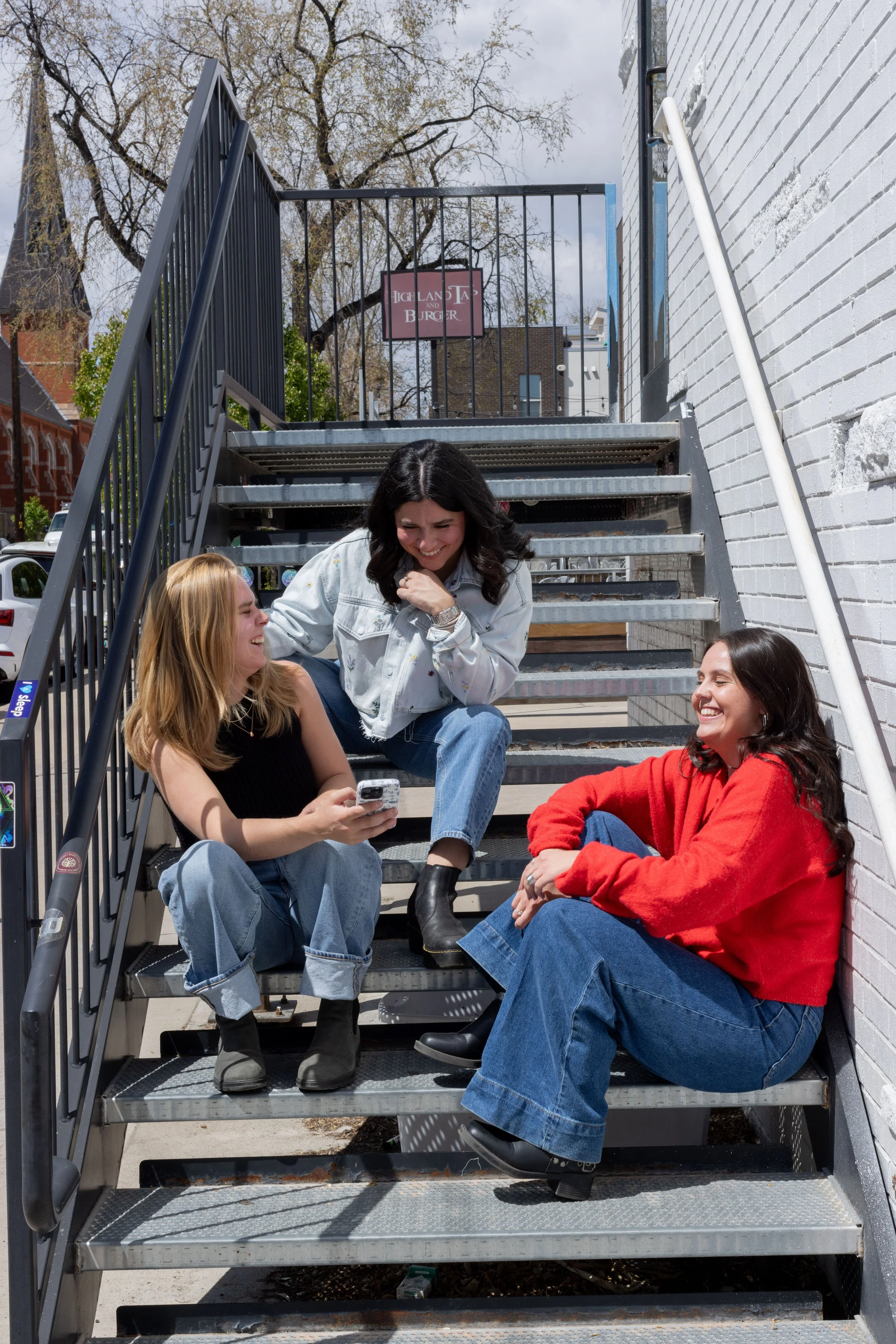 Cucamonga Media team members Casey, Alix, and Lauren outside sitting on steps laughing and playing on their phones
