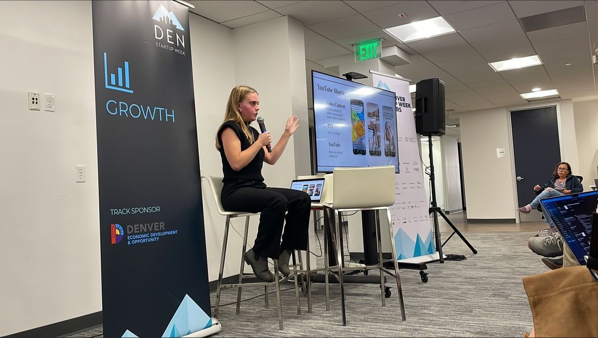A woman is giving a presentation at a conference, seated on a high stool, holding a microphone, with a large screen displaying slides behind her. The setting is a conference room with an audience, banners, and a visible exit sign.
