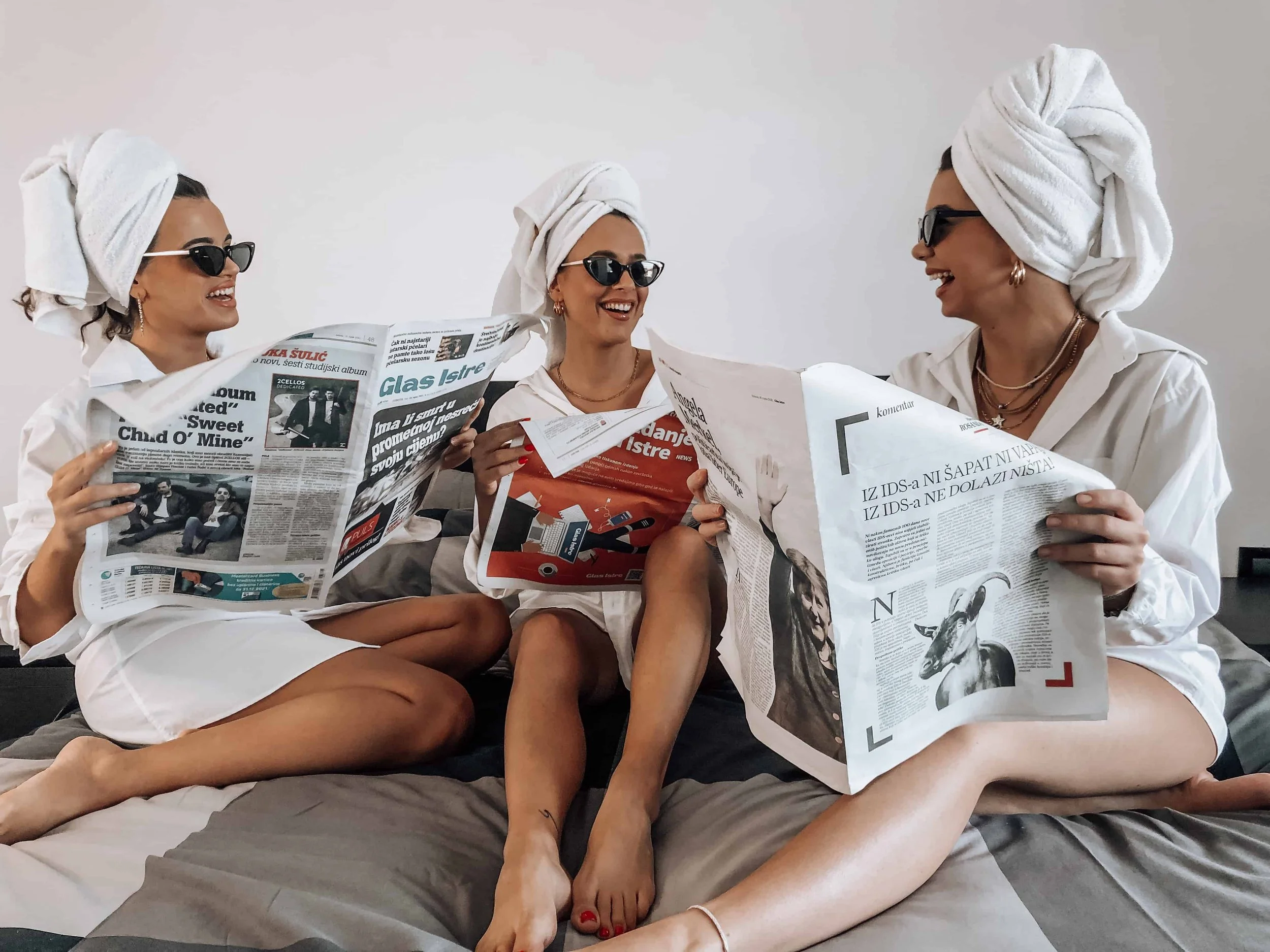 Three women in white shirts, towel turbans, and sunglasses sit on a bed laughing together while reading newspapers in a playful, relaxed morning scene.