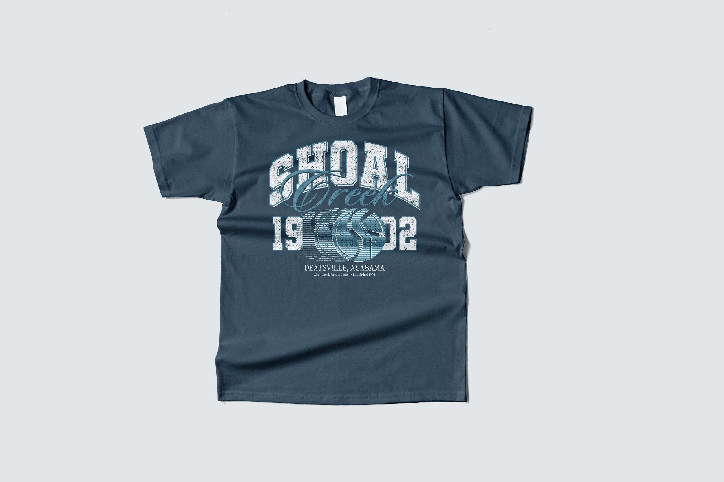A blue T-shirt with white and light blue printed text that says 'SHOAL Creek 1992' and 'Deatsville, Alabama' along with a graphic of a stream or river.
