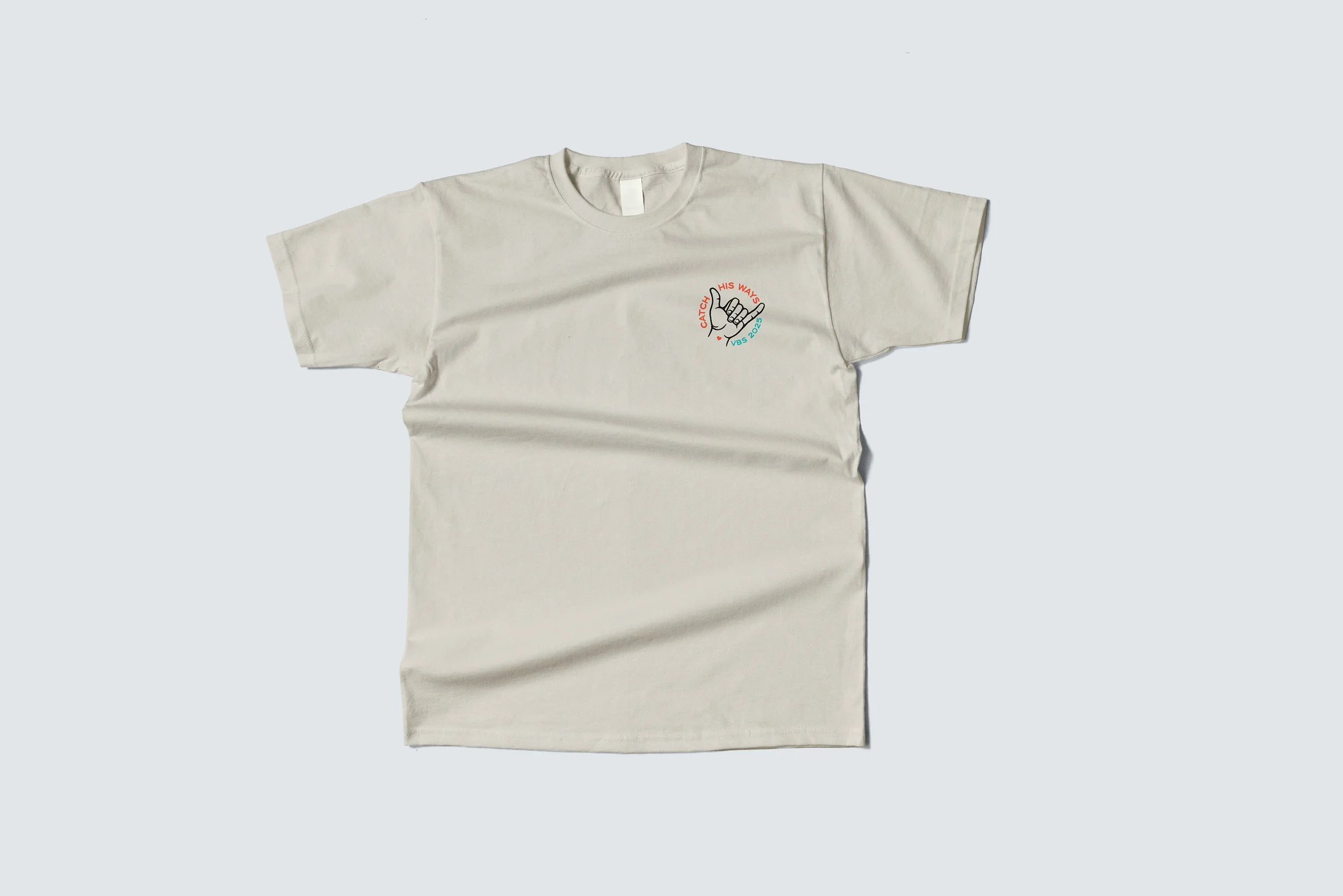 White T-shirt with a pocket on the left chest displaying a small graphic of a hand making a thumbs-up gesture, surrounded by red, black, and teal text that reads 'Catch His WAVE! NYS 2023'.