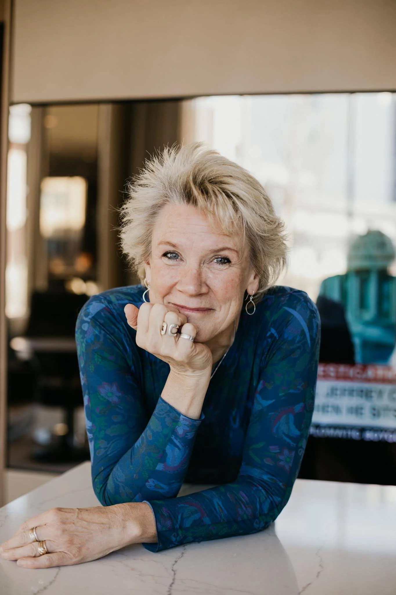 Karen leans on a table in a long-sleeve blue shirt, smiling at camera.