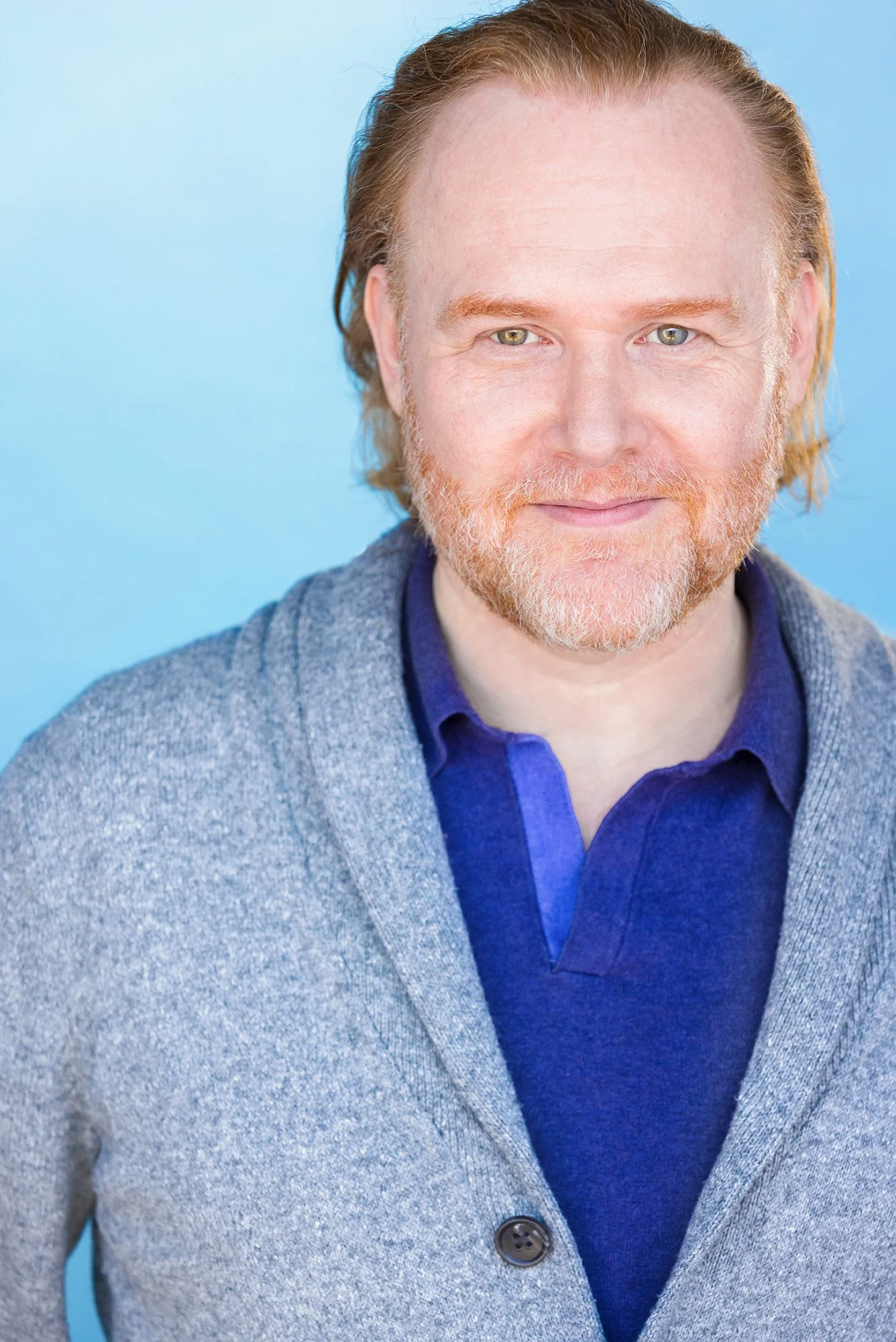 John Flynn has red hair and wears blue shirt and gray cardigan.