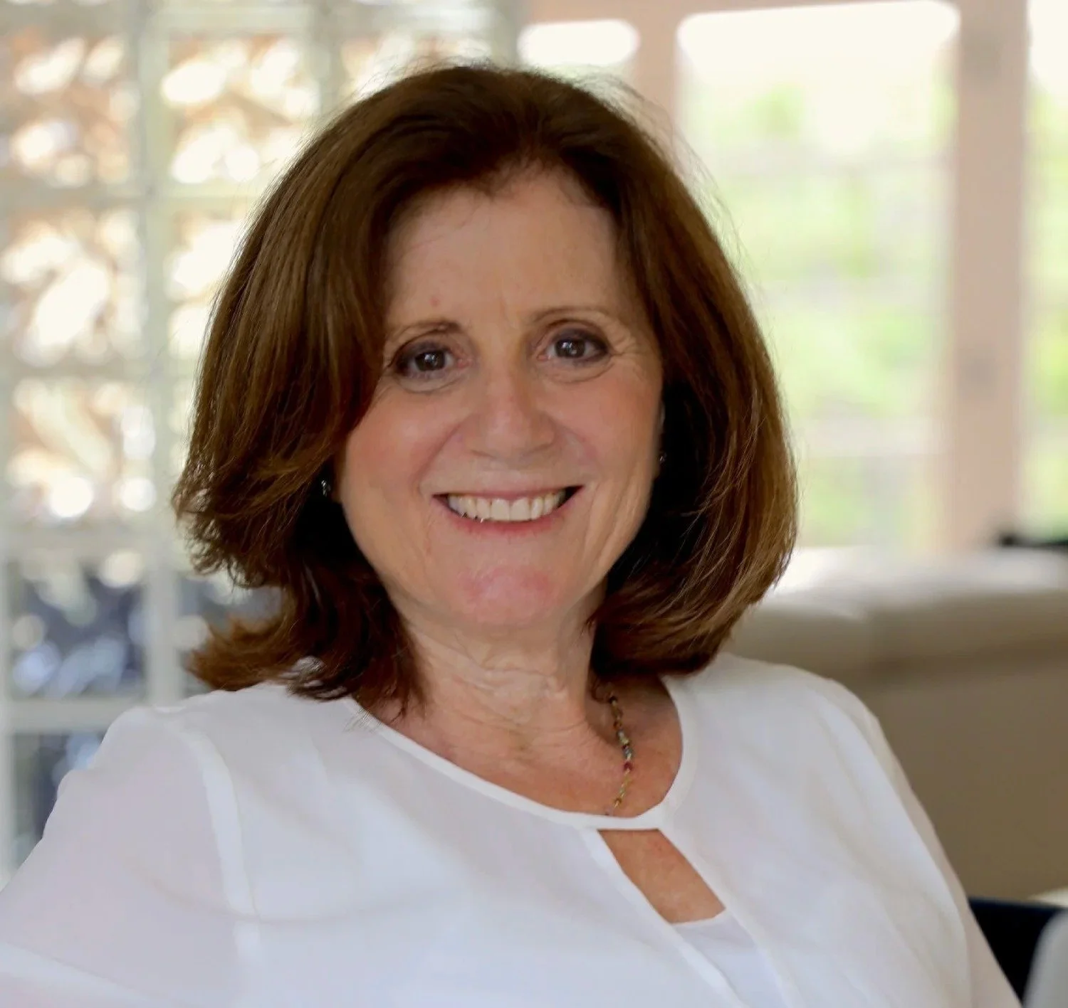Close-up of a smiling middle-aged woman with shoulder-length brown hair, wearing a white top, sitting indoors in front of a window with blurred greenery outside.