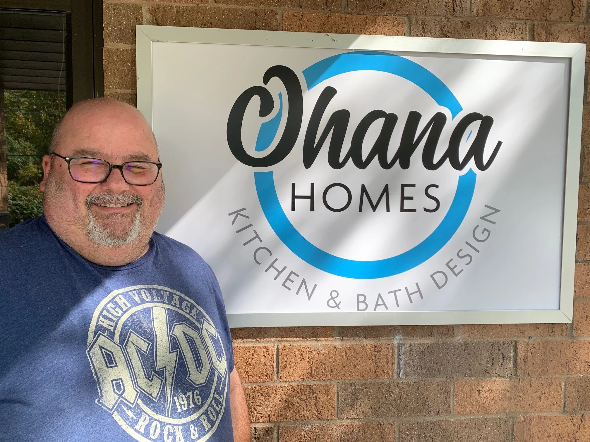 A smiling man with glasses and a beard standing outside next to a sign that reads 'Ohana Homes Kitchen & Bath Design' against a brick wall.