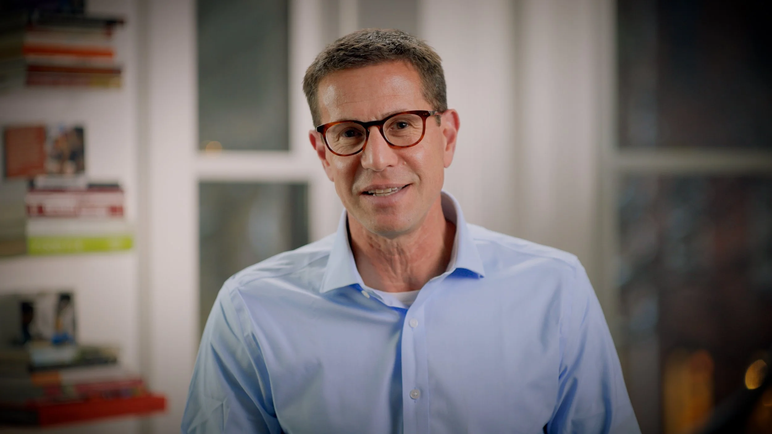 A man with brown hair and glasses wearing a light blue dress shirt, standing in a room with bookshelves and windows in the background.