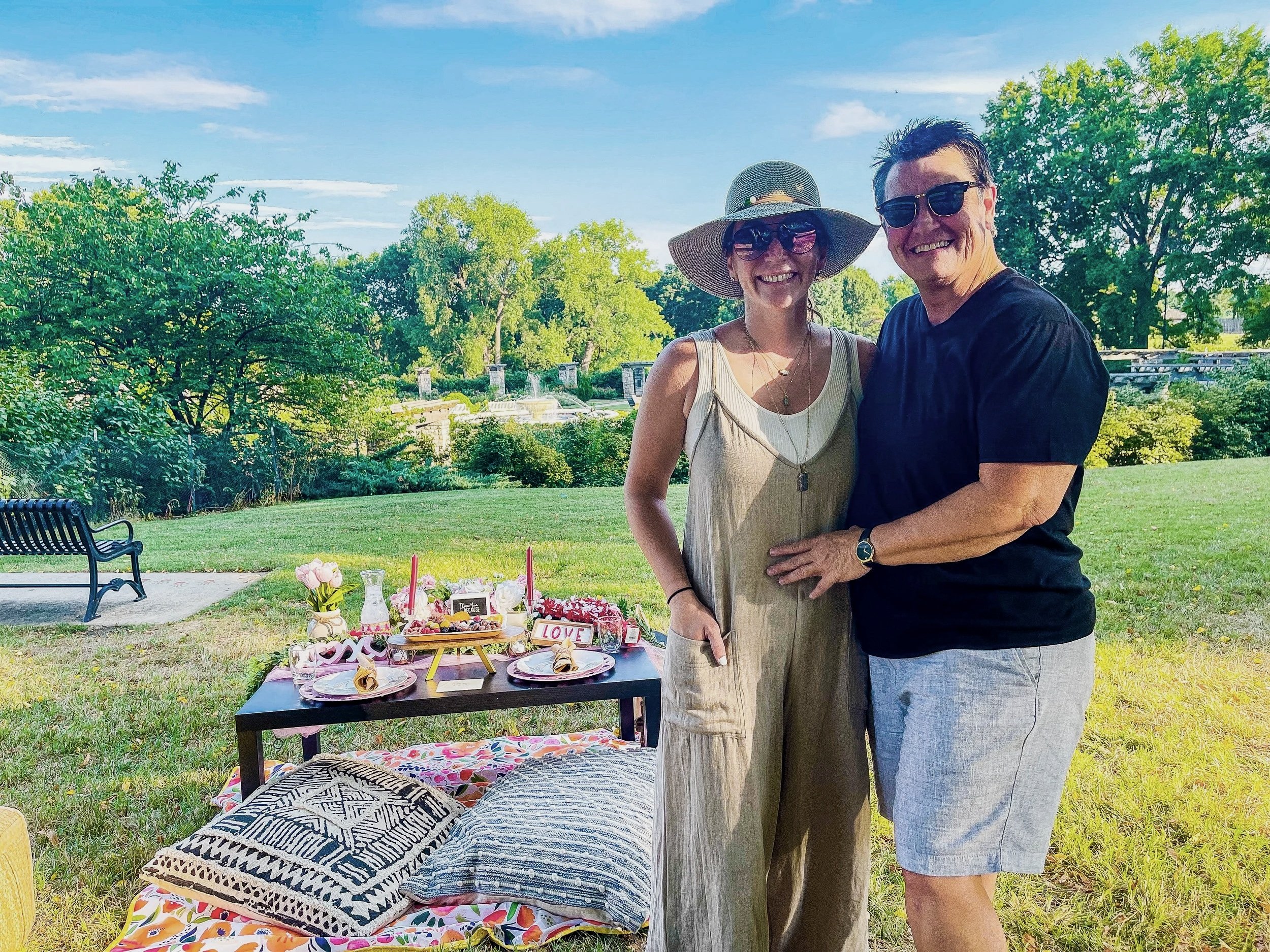 A smiling woman and man standing outdoors at a garden party, with tables set with flowers and decorations, surrounded by green trees and blue sky.