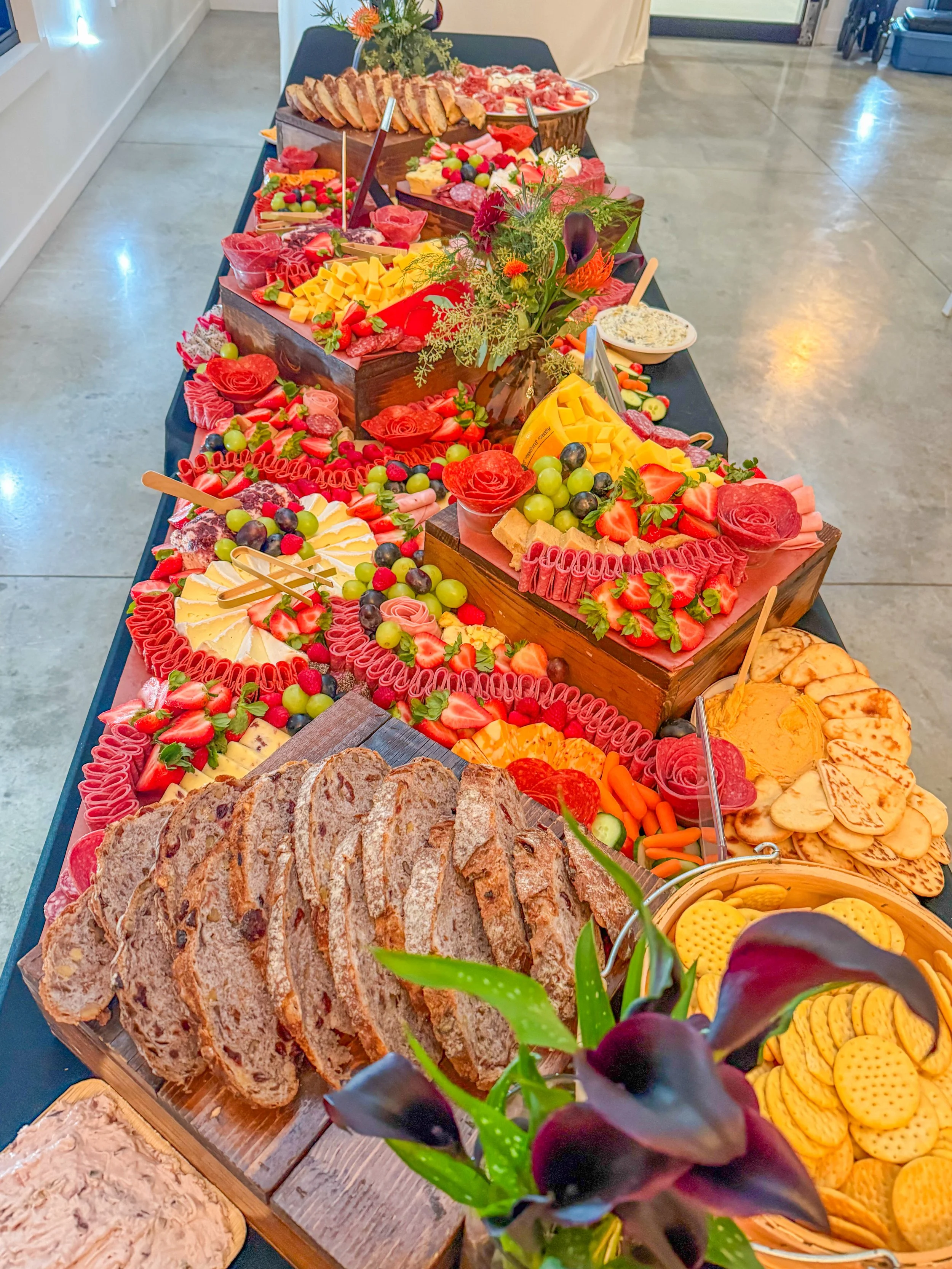 A long table with a variety of colorful food and desserts such as sliced bread, cheese, strawberries, grapes, cookies, and a cheese platter, decorated with flowers.