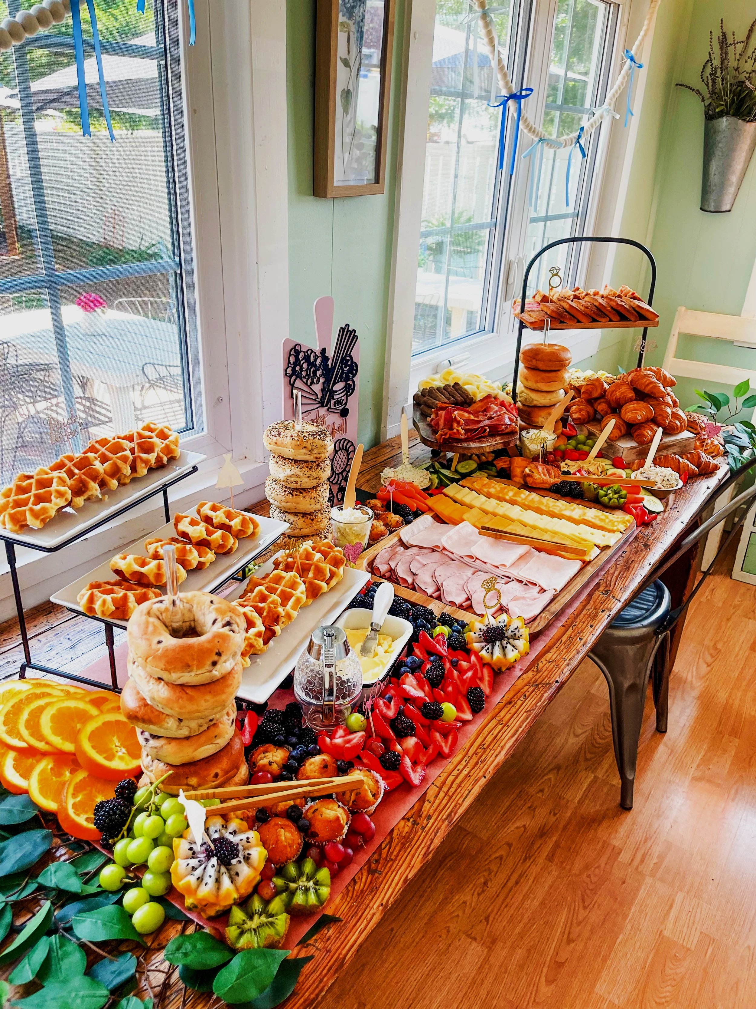 A table filled with breakfast foods including waffles, bagels, bagel chips, sliced oranges, grapes, strawberries, blackberries, and various cheeses, with a view of a bright room with large windows.