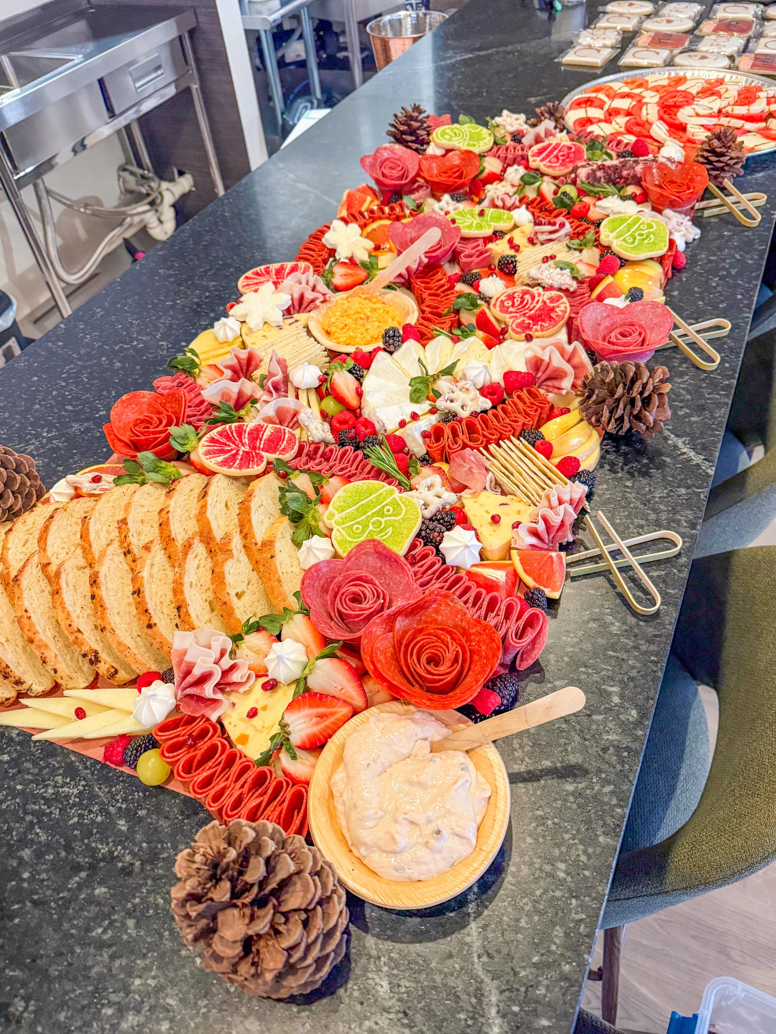 Colorful holiday cheese and meat platter with sliced bread, strawberries, raspberries, and decorative pinecones on a dark countertop.
