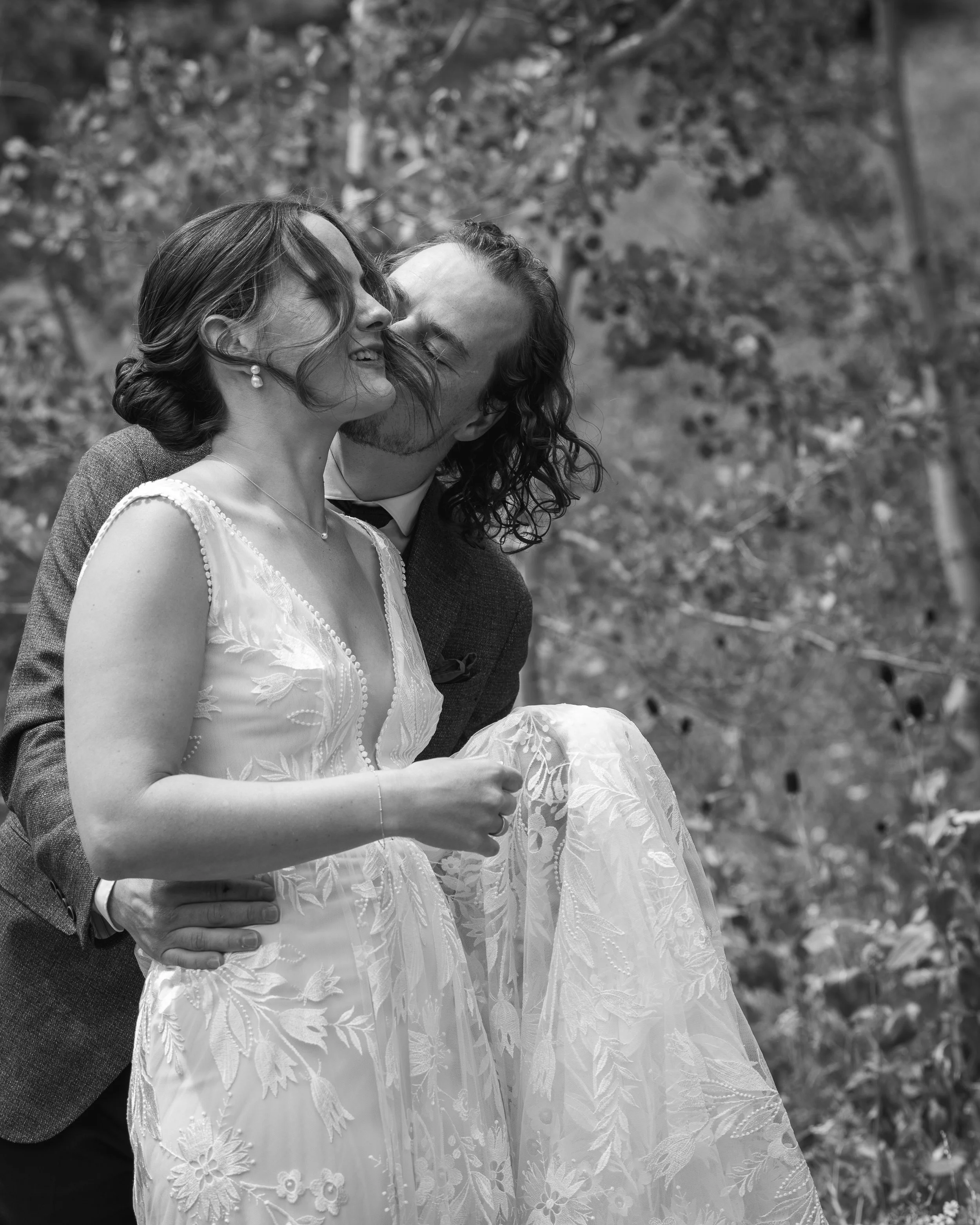 Black and white documentary style wedding photography, showing groom behind bride, holding her waist, kissing her cheek