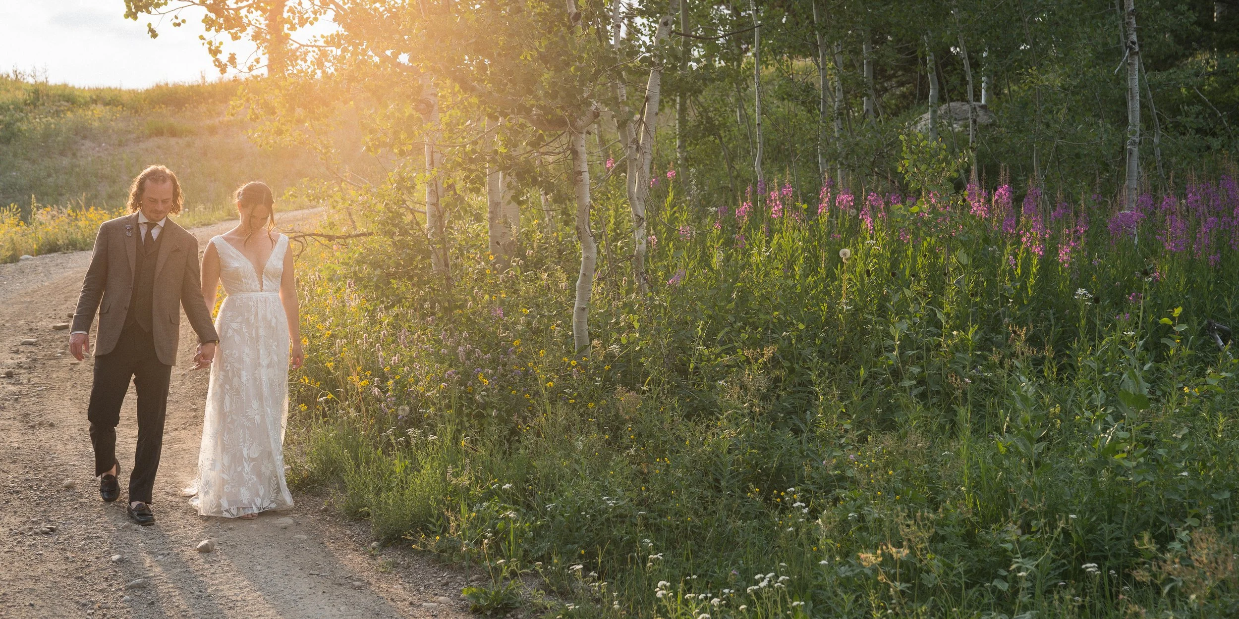 Grand Targhee Resort, documentary Wedding photography; bride and groom walking toward camera with sun in background, illuminating purple fireweed flowers