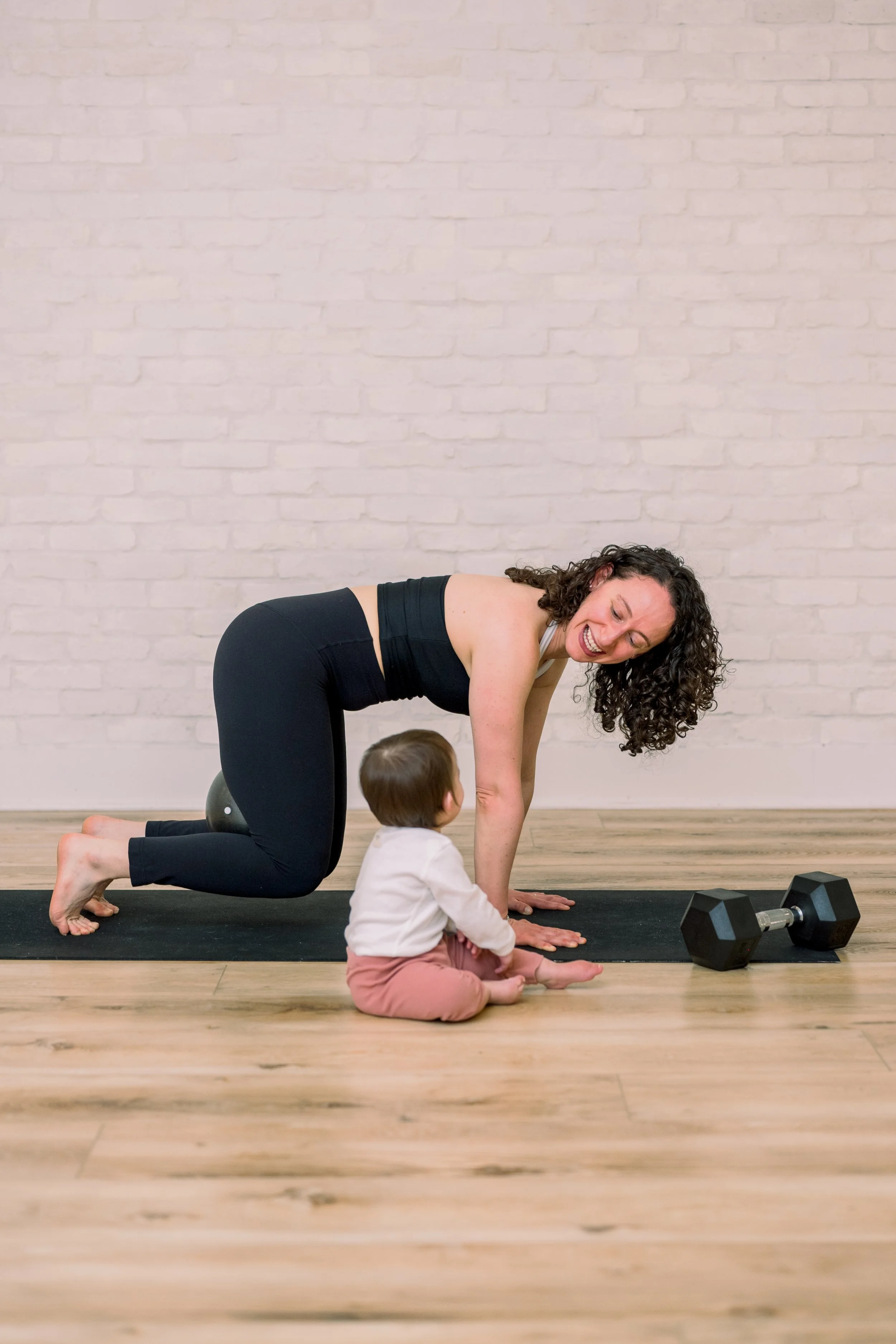 A woman practicing yoga indoors, raising a weight over her head, standing in a lunge position with one hand on her stomach, wearing a green sports bra and green leggings.