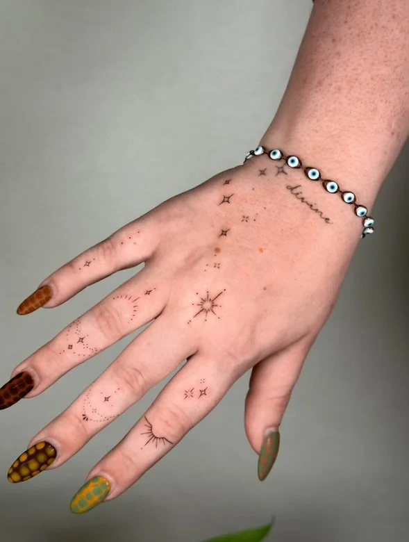 Close-up of a hand with small tattoos, including stars and a compass, and colorful patterned nails. The person is wearing a bracelet made of eyeball beads and has a tattoo that reads 'using' on the wrist.