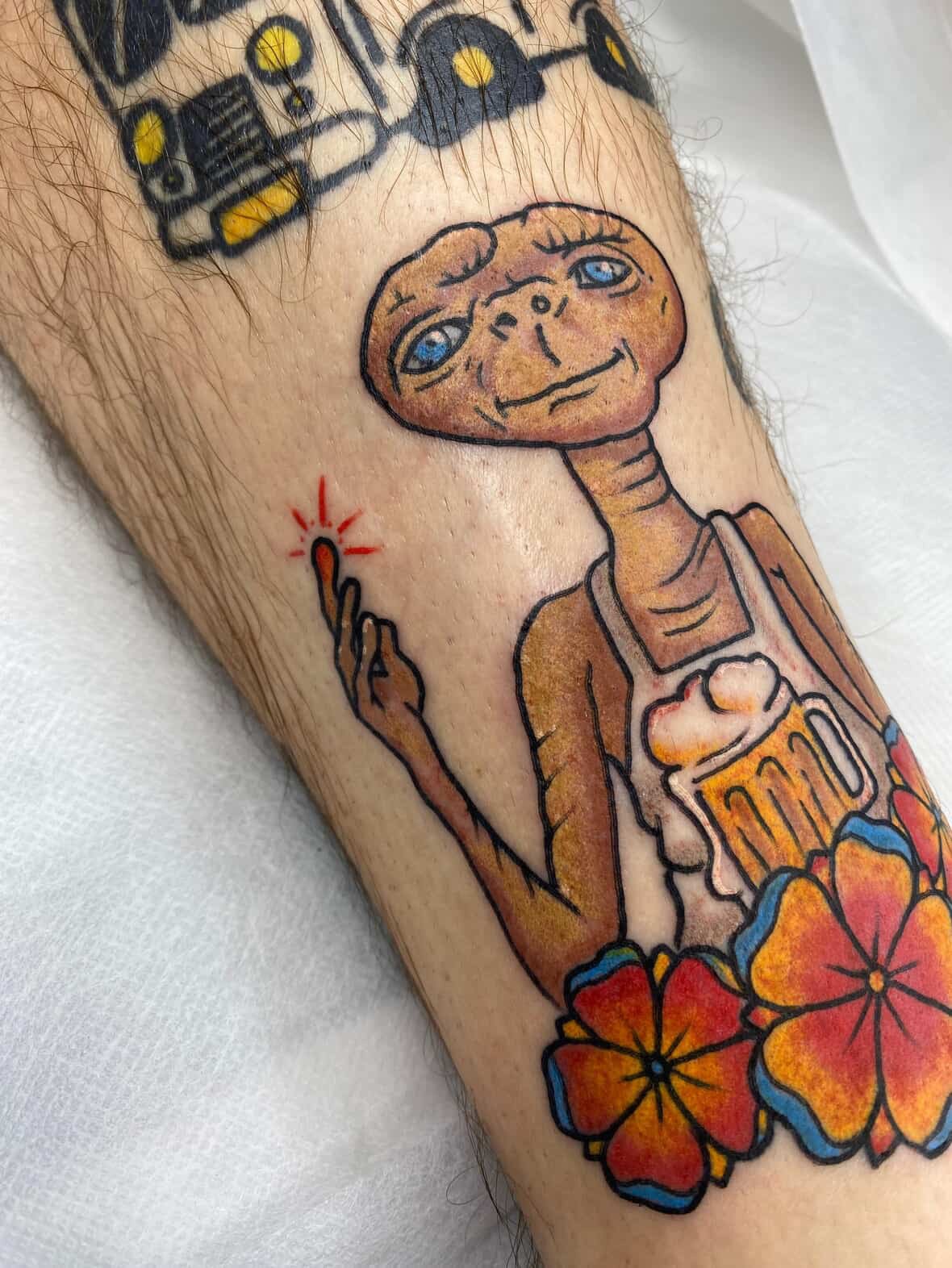 Tattoo of E.T. holding a beer mug, with colorful flowers below, and part of a mechanical robot in the background.