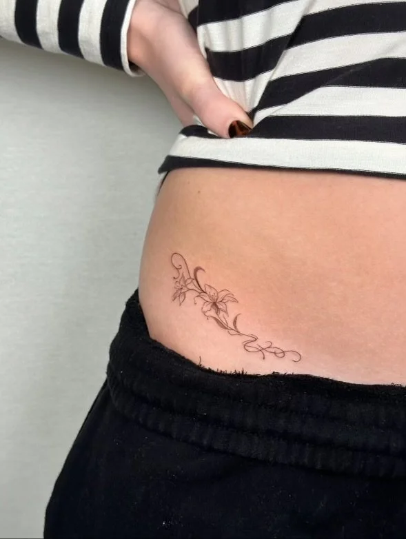 Tattoo of a floral vine with leaves and swirls on the person's side near the hip.