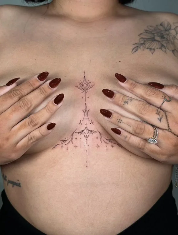 A person with tattoos on their chest and hands holding their breasts. The tattoo on their chest appears decorative and symmetrical, with fine line details.