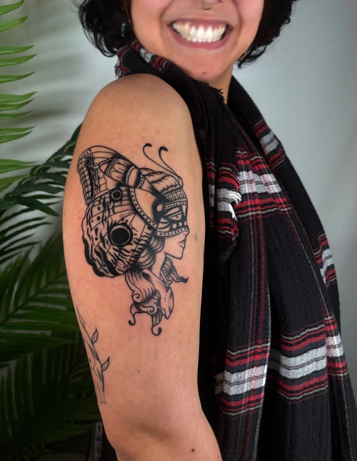 Tattoo of a woman's profile with a headdress on the upper arm of a smiling woman with dark hair, wearing a black and red plaid scarf, standing near green plant leaves.