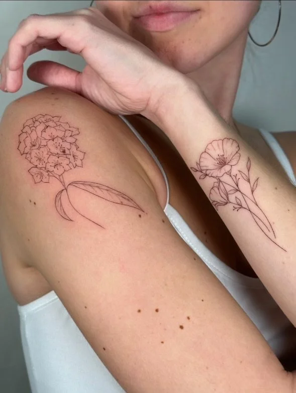 A woman shows her shoulder and forearm tattoos of delicate floral designs, one on her shoulder and one on her forearm, both in a minimalist line art style.