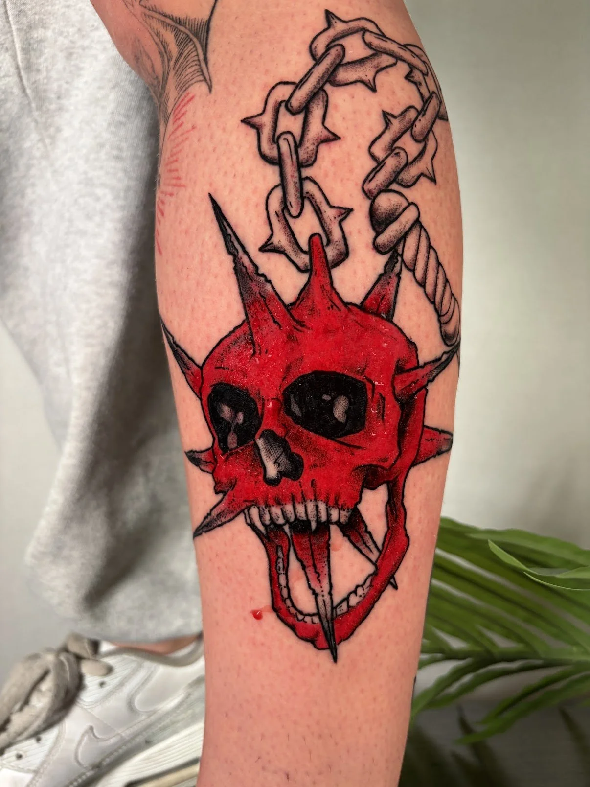Tattoo of a red skull with black eye sockets and nose cavity, dripping with red ink, surrounded by a jagged starburst shape and attached to a chain. The skull has spikes protruding and a large needle through the lower jaw.