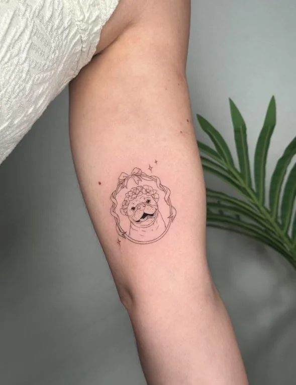 Tattoo of a smiling dog with a flower crown and wavy hair, surrounded by an oval frame and small sparkles, on person's upper arm.