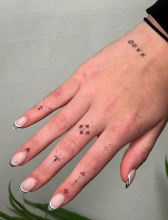 A person's hand with small, minimalist tattoo designs on fingers and back of the hand, including symbols and geometric patterns.