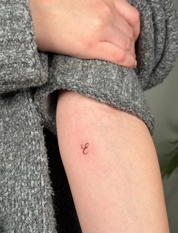Close-up of a person's upper arm with a small, minimalist tattoo of a cursive letter 'E' on the inner forearm, partly covered by a gray, textured sweater.