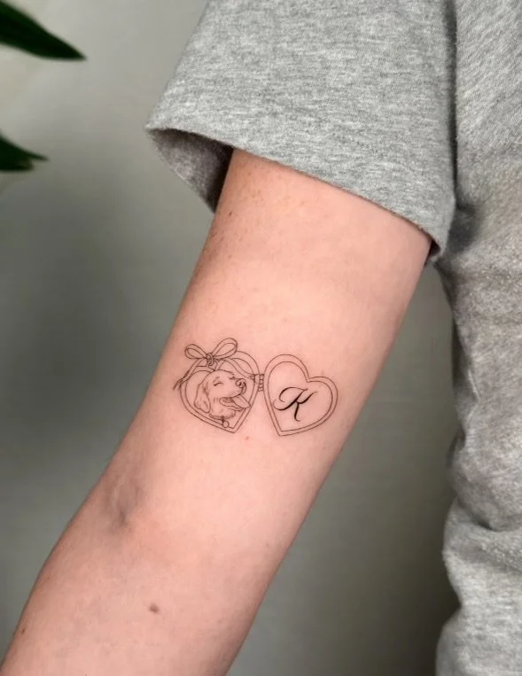 Tattoo of two linked hearts on a person's arm; one heart has a drawing of a girl with a bow in her hair, and the other contains the letter 'K'.