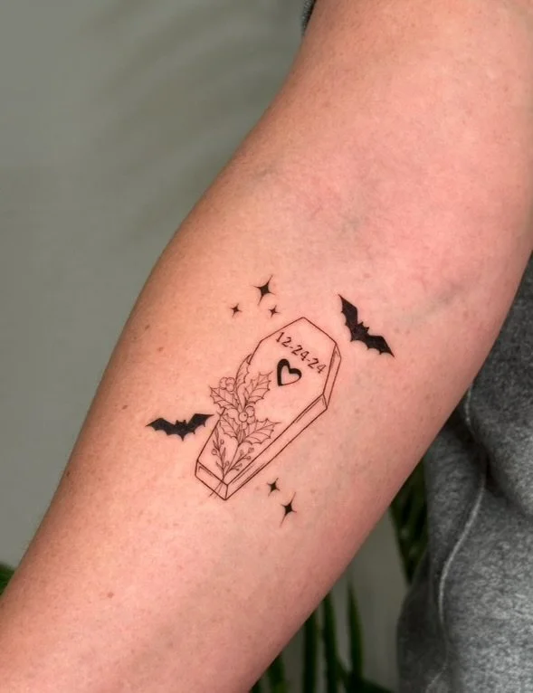 Tattoo of a coffin with the date 12-24-24 and a heart on it, surrounded by holly leaves, berries, and three small stars, with two bats flying nearby.