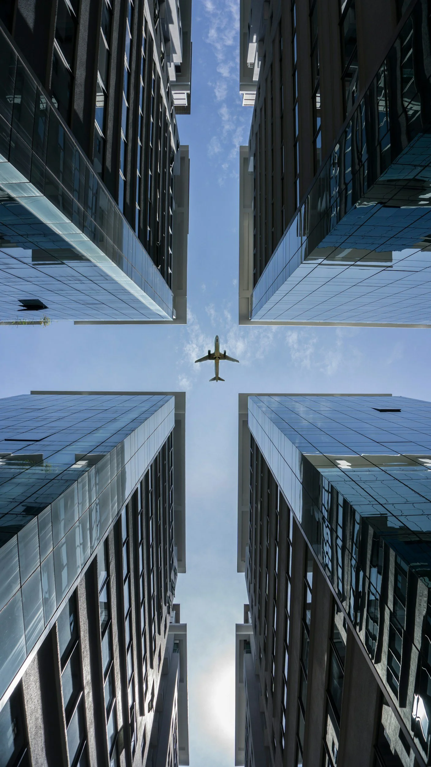 Looking up at the sky through a narrow gap between tall glass buildings, with an airplane flying overhead.