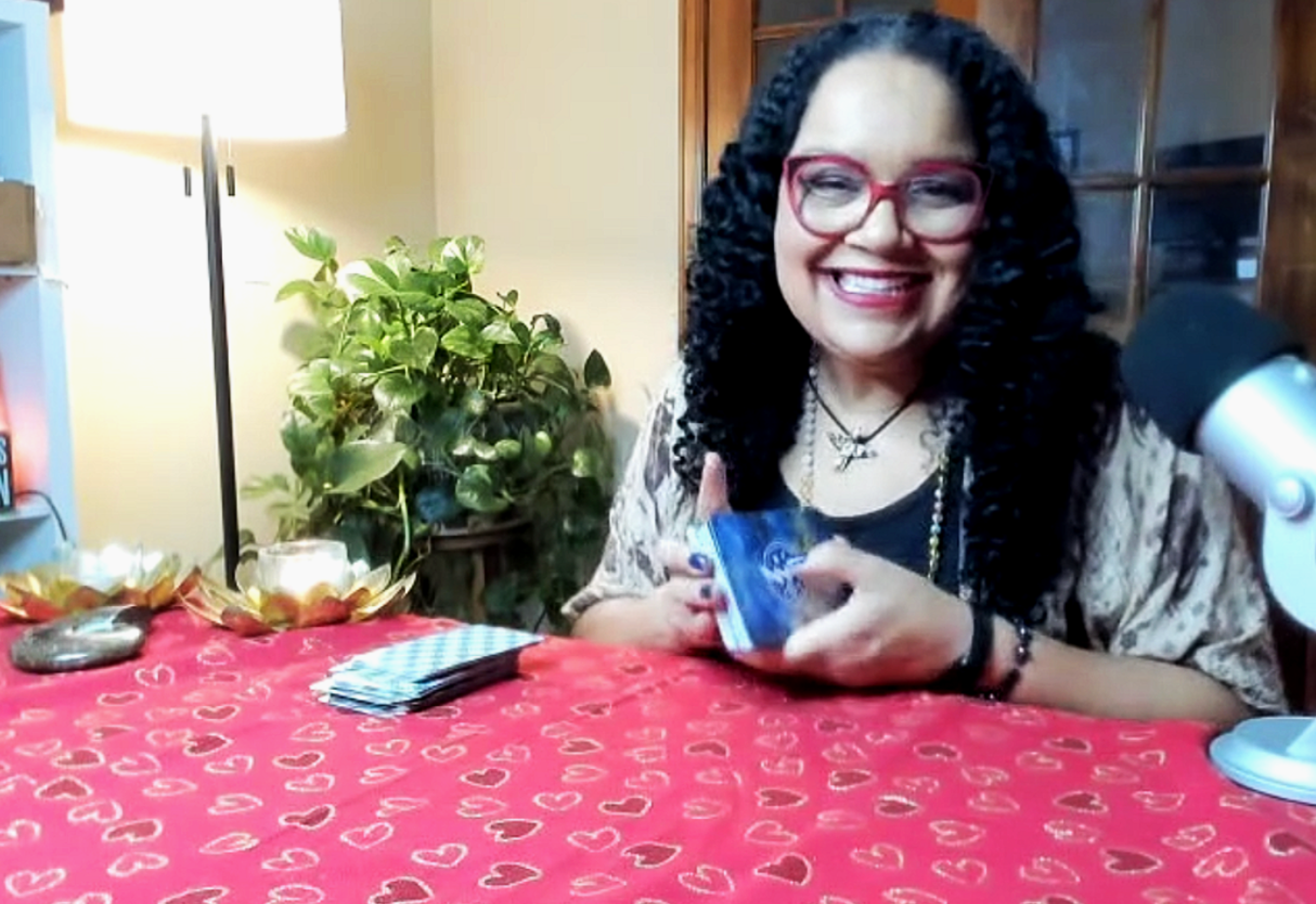 A woman with curly black hair, red glasses, and a bright smile, sitting at a table with a red tablecloth decorated with white hearts. She is holding a deck of cards, with a microphone pointed towards her, in a cozy indoor setting with a lamp, green plant, and wooden cabinet in the background.
