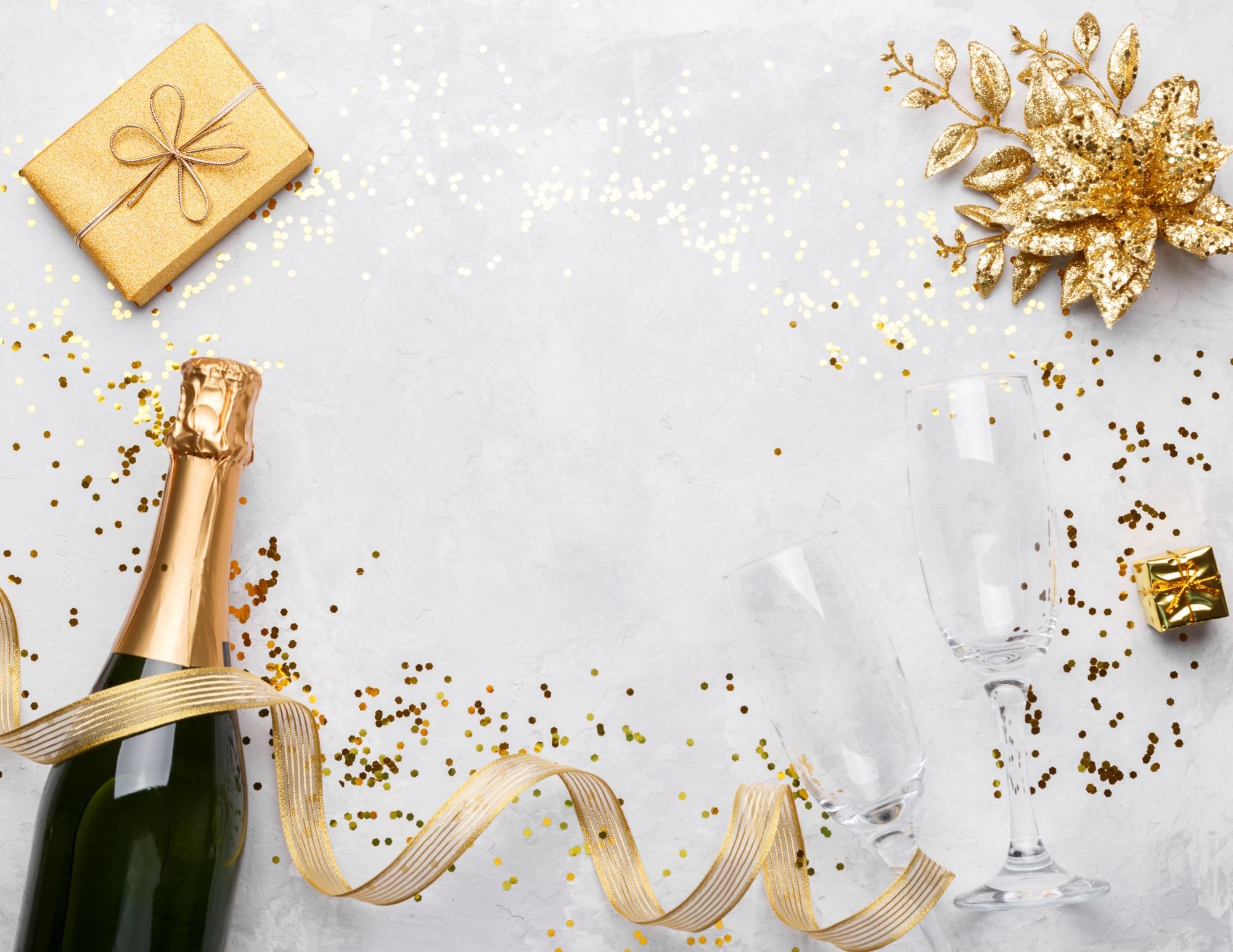 Gold Christmas decorations, champagne bottle, champagne glasses, small gift box, and gold confetti on a white surface for celebration.
