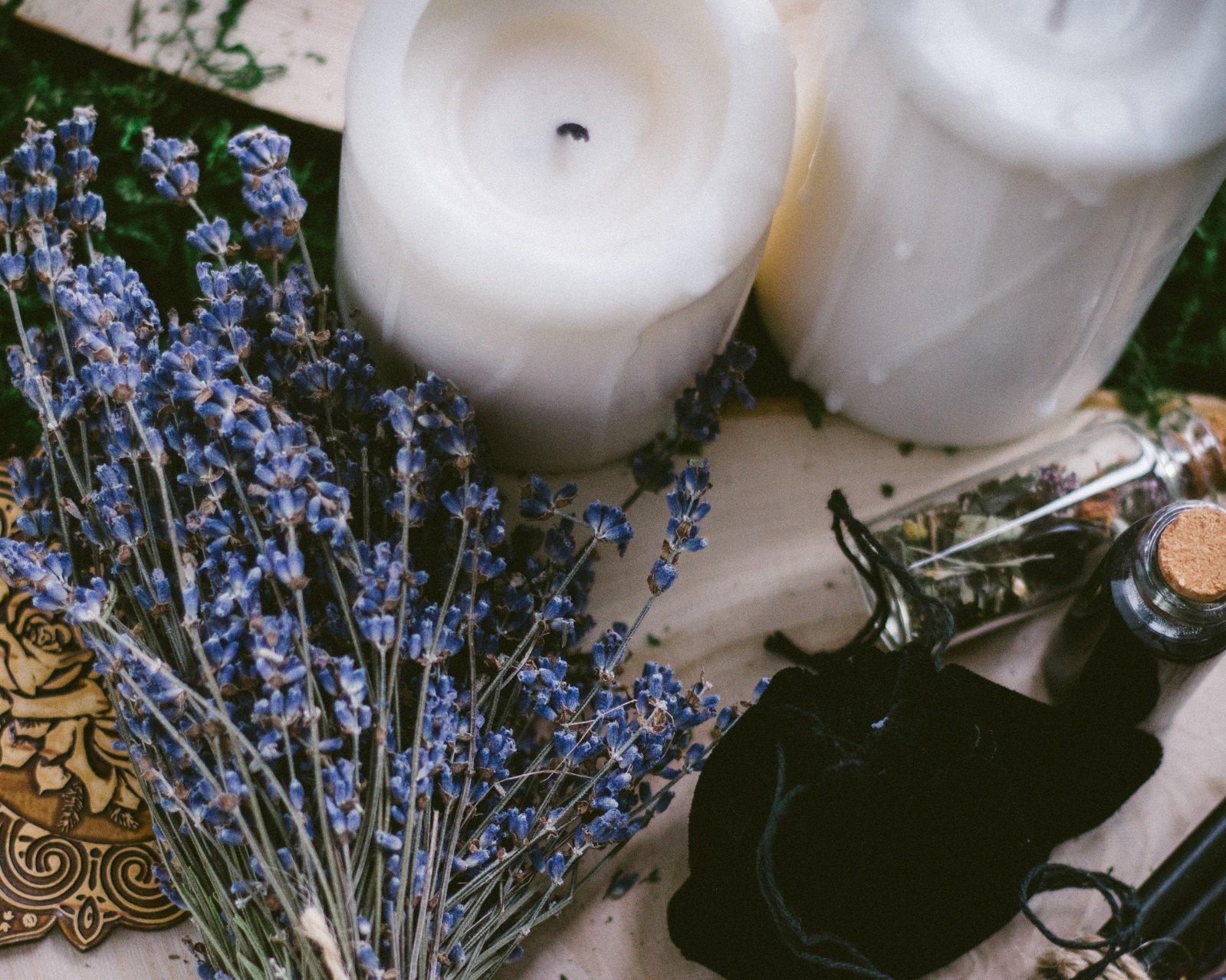 A collection of items including a bundle of dried lavender, white candles, a glass jar with dried herbs, a small bottle with a cork stopper, and black string or cloth on a wooden surface.