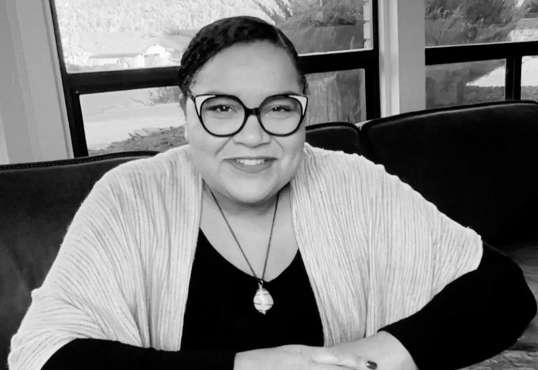 A smiling person with short dark hair, glasses, and a necklace, sitting at a table in a room with large windows in the background.
