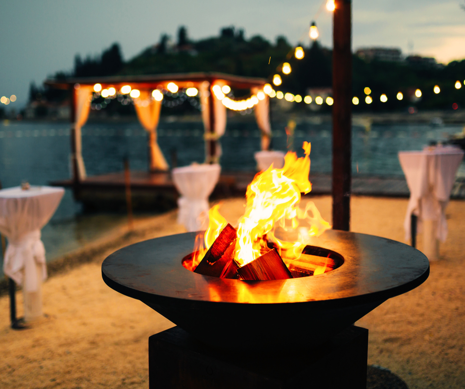 A bonfire on a beach at dusk with a lakeside pavilion decorated with string lights in the background.