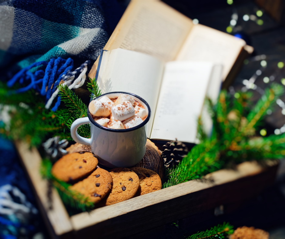 A holiday-themed setup with a mug of hot chocolate topped with marshmallows, surrounded by chocolate chip cookies, pinecones, and evergreen branches, with an open book and a blanket in the background.