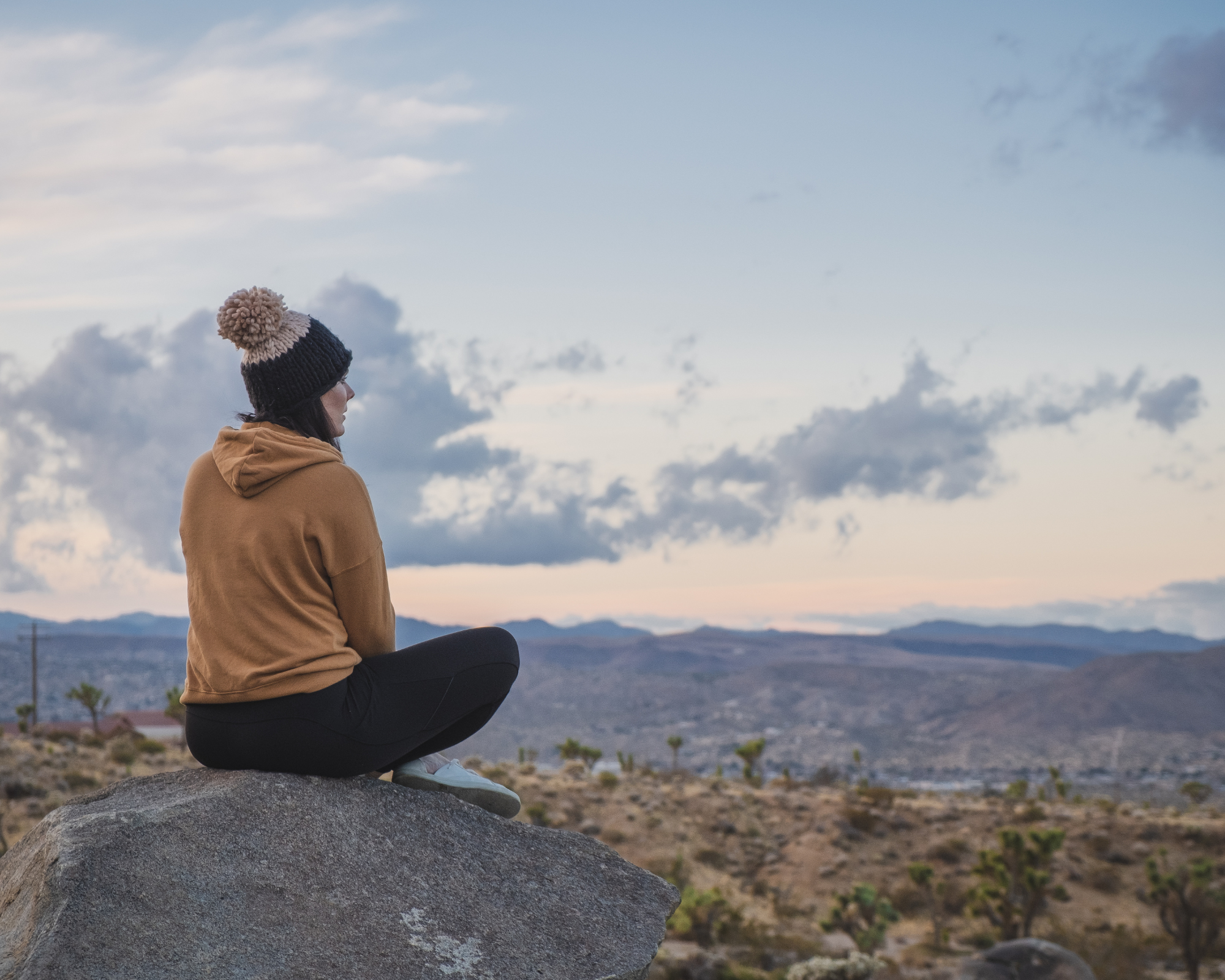 A woman sitting on a rock high above a city looking out over the distance as the sun sets.