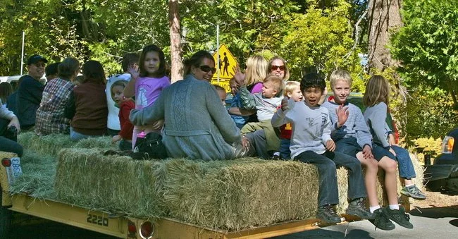 Children and adults sit on hay bales on an outdoor hayride trailer beneath trees.